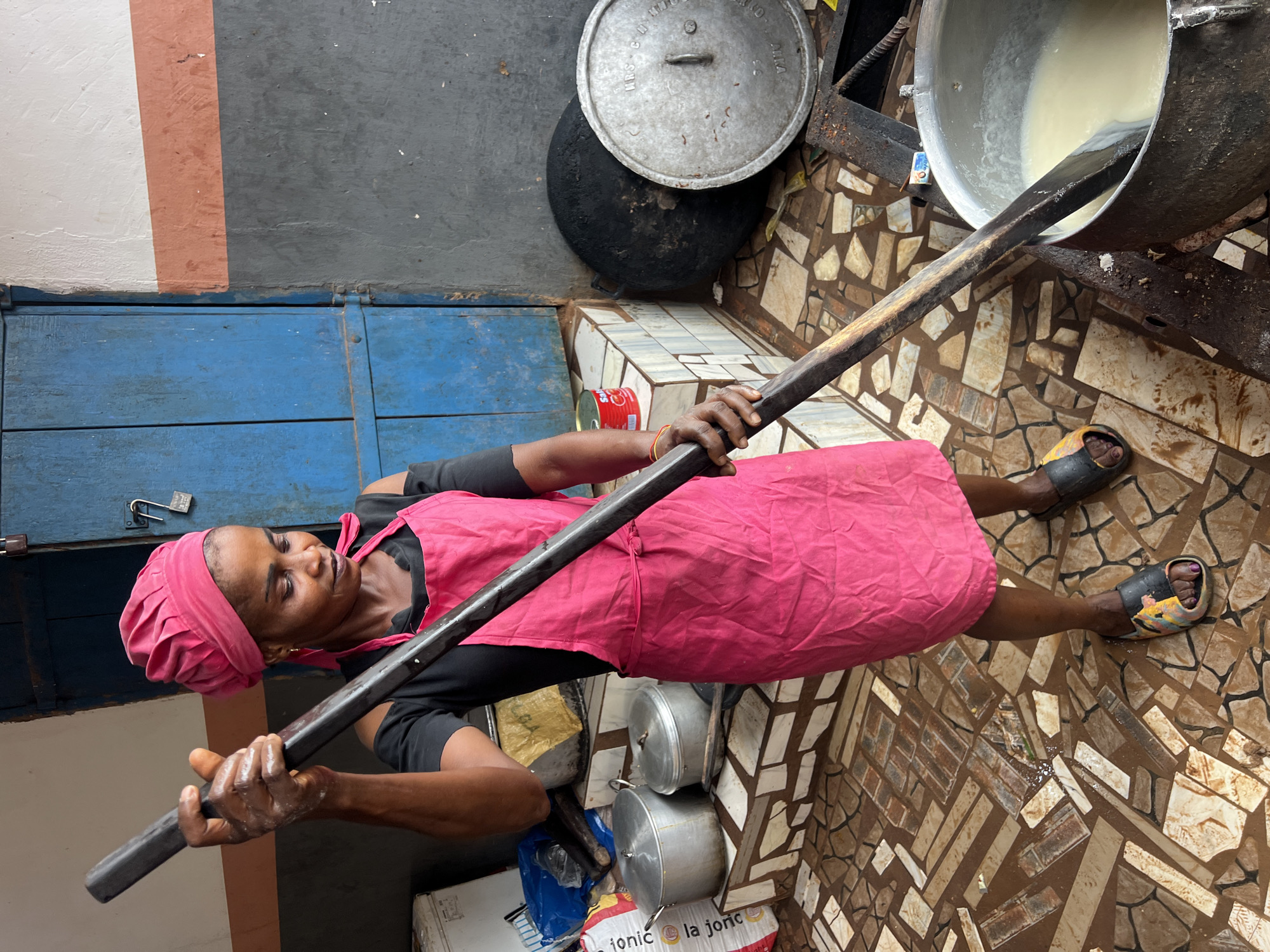 In Nkpor, Nigeria, a woman wearing a pink headscarf and apron stirs a pot of light-colored liquid with a large wooden spoon. She is standing in a cluttered outdoor kitchen area with a tiled floor. To her left are several large metal pots and bags of supplies. To her right, a large black wok-like pot sits next to a lidded pot. A blue wooden door with two locks is visible in the background. The overall scene depicts food preparation in a busy, possibly commercial, setting.