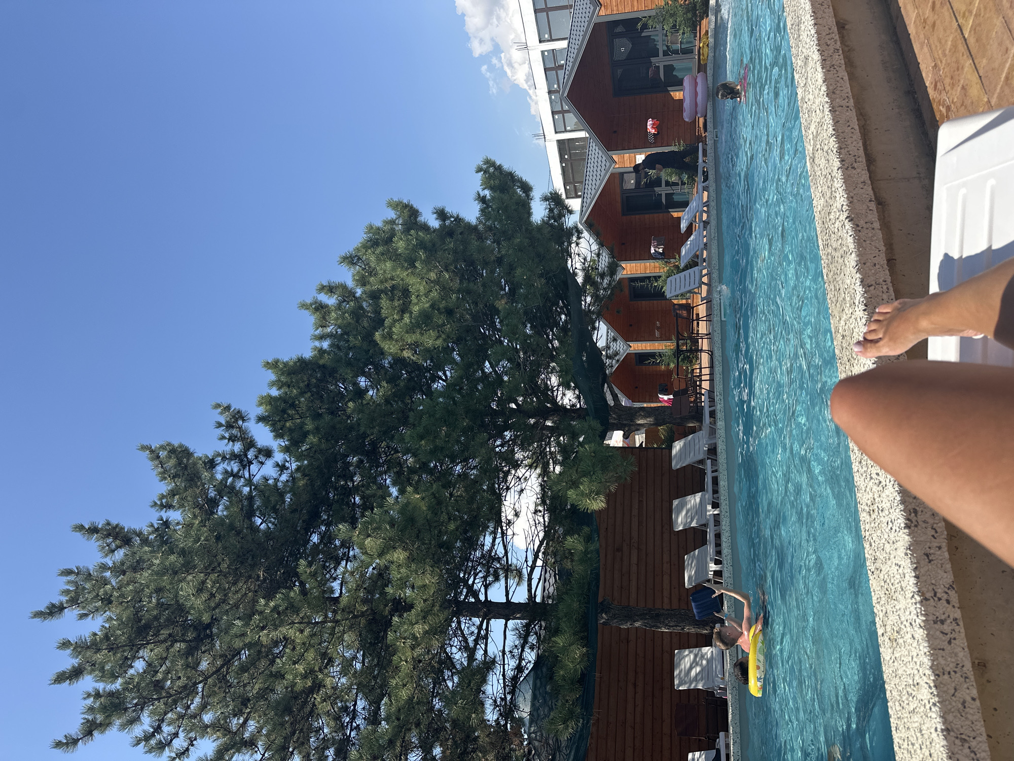 The image depicts a poolside scene at a resort in Anapa, Russia. A clear blue swimming pool occupies the foreground, with a person's legs visible from a reclining position at the bottom of the frame. Two children are visible in the pool; one boy is on a yellow inflatable ring, holding a dark blue object, and a girl is further back, partially submerged. In the background, several identical wooden cabins with dark roofs are lined up, suggesting a holiday village or motel. A large, mature pine tree with dense green foliage stands prominently on the left, casting shadows. Lounge chairs are arranged by the pool and in front of the cabins. The sky is clear and blue, with a few wispy clouds visible.