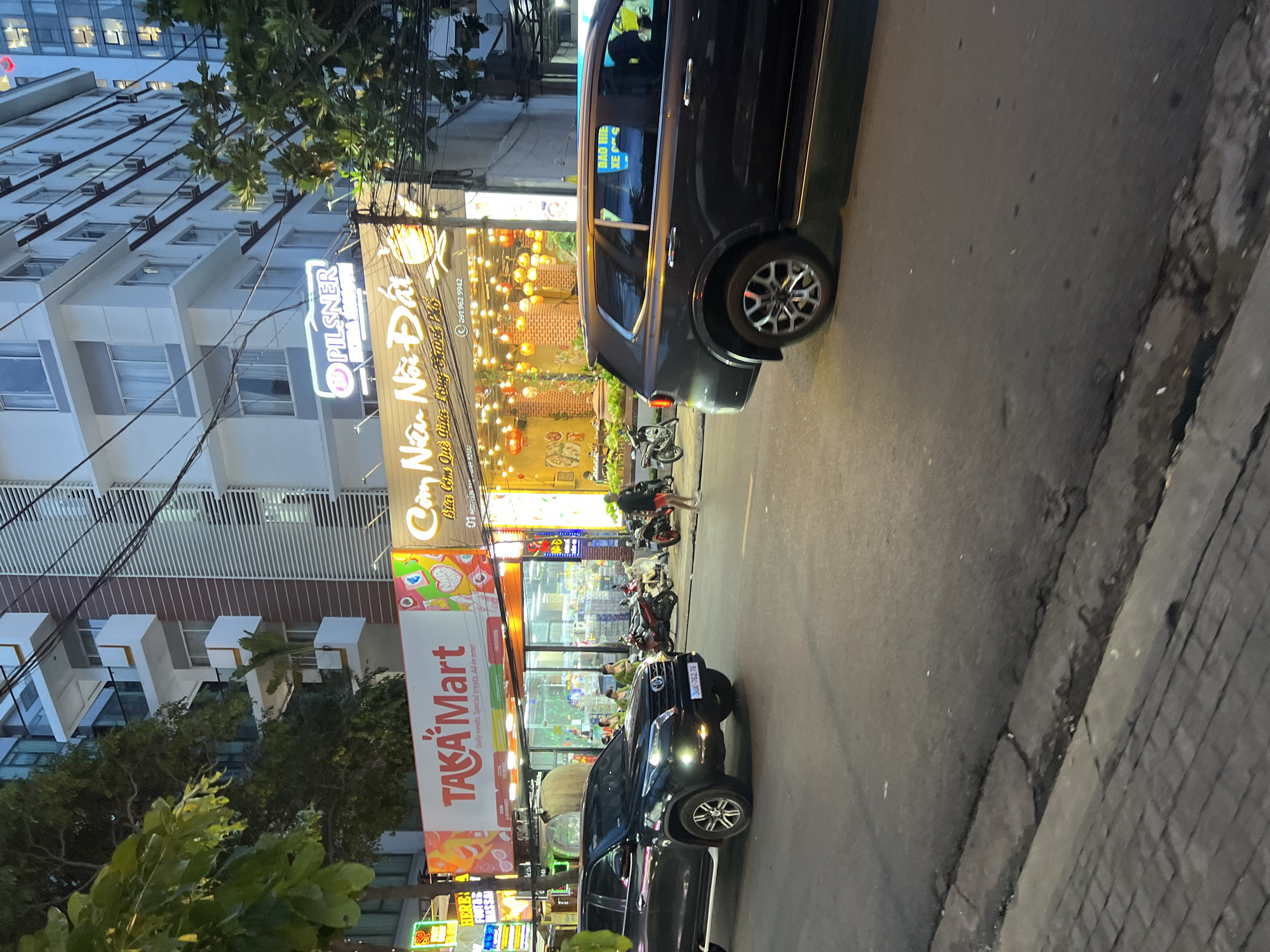 The image captures a street scene in Nha Trang, Vietnam, likely in the evening. In the foreground, a dark-colored Toyota SUV is driving on the right side of the road. To the left, a black Toyota Fortuner SUV is parked on the street. Behind the vehicles, a row of commercial establishments lines the sidewalk. The most prominent is "TAKA Mart," a convenience store with a bright red and white sign, advertising "Daily needs. Special treats. All in one!". Next to it, a restaurant called "Cơm Nhà Nổi Đạt" features a brightly lit interior with numerous hanging lanterns and a sign advertising a phone number. Several motorcycles are parked in front of the establishments, and a few individuals are visible on the sidewalk, appearing to be patrons or employees. The background reveals a modern multi-story building with balconies and visible air conditioning units, suggesting an urban environment. The overall lighting indicates it is either dusk or the streetlights and business signs are illuminated. The road surface is asphalt, and a paved sidewalk with a concrete curb is visible.