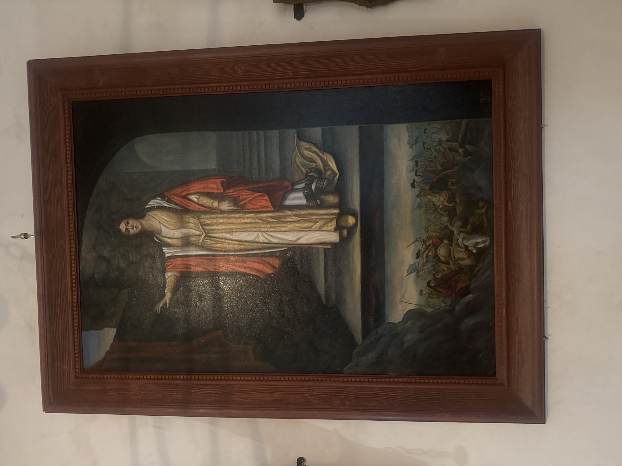 Here is a description of the image:
A framed painting hangs on a white wall. The painting depicts a person lying on a bed with a red garment draped over them. A group of figures, possibly soldiers or warriors, are depicted in the background on the right side of the painting. The environment appears to be a classical setting, with architectural elements visible. The location is the town of Chynadiiovo, Ukraine.
