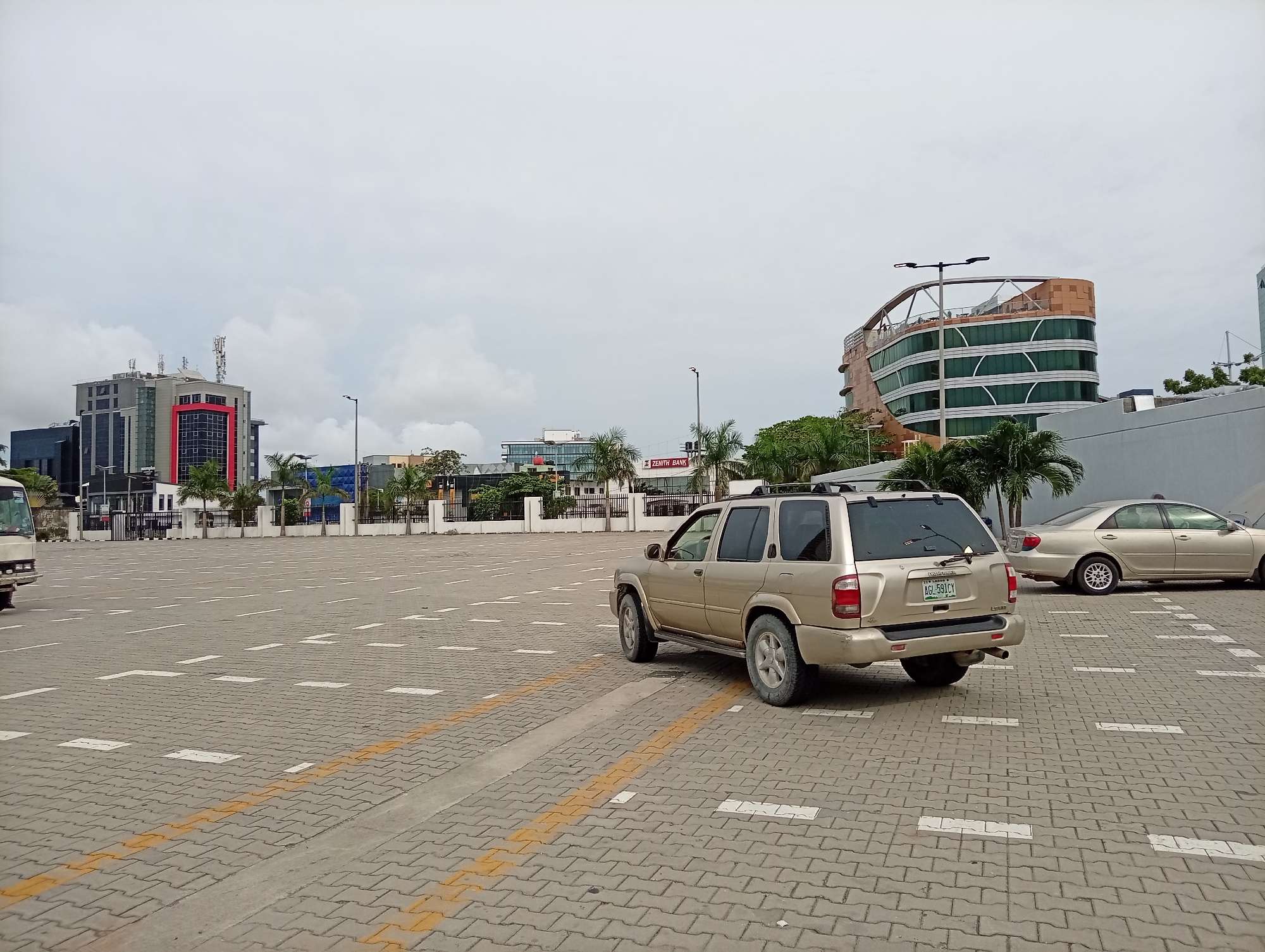 The image shows an outdoor parking area in Itirin, Nigeria, under a cloudy sky. The parking surface is paved with gray interlocking blocks marked with dashed white parking lines and solid yellow lines. A gold Nissan Pathfinder SUV, facing away from the camera, is in the parking area. To the left of the SUV is a partial view of a white van or bus. To the right, a gold sedan is parked. In the background, several modern buildings can be seen, including a building with a red accent and antennae on top, a building with a curved design, and a Zenith Bank building. Palm trees are scattered among the buildings.