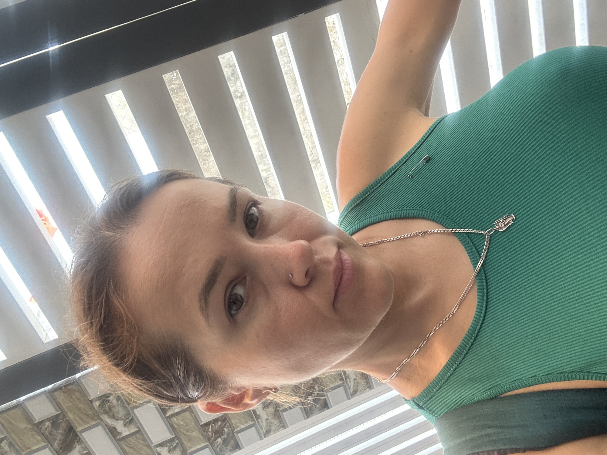 A woman is the primary subject of the image, captured from a low angle. She has fair skin, brown hair, and light-colored eyes, with a visible nose piercing. She's wearing a green ribbed tank top and a silver necklace. The background includes light-colored blinds and tiled flooring. No specific details connect the scene to Bac Nha Trang Ward, Vietnam.