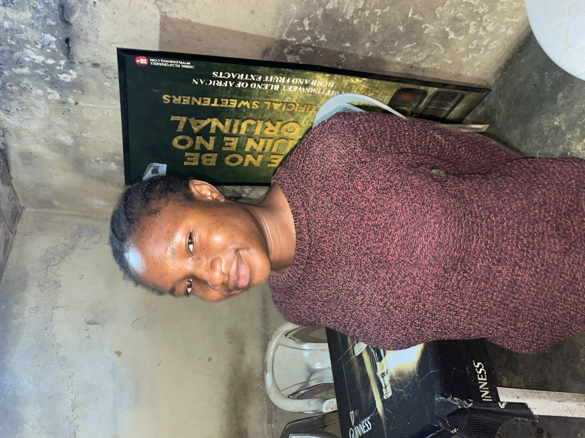 A young Black woman is centered in the frame, her head and shoulders visible. She has dark hair pulled back, dark eyes, and a warm complexion. She wears a dark pink knit sweater. Behind her is a worn wall with a dark green advertisement, partly obscured, with the words "BE ORIGINAL," "SWEETENERS," and "AFRICAN HERB AND FRUIT EXTRACTS" visible. A black box with "GUINNESS" written on it is partially visible. The environment appears to be an indoor space with concrete floors and walls. The location within Jos, Nigeria, cannot be determined from the image.