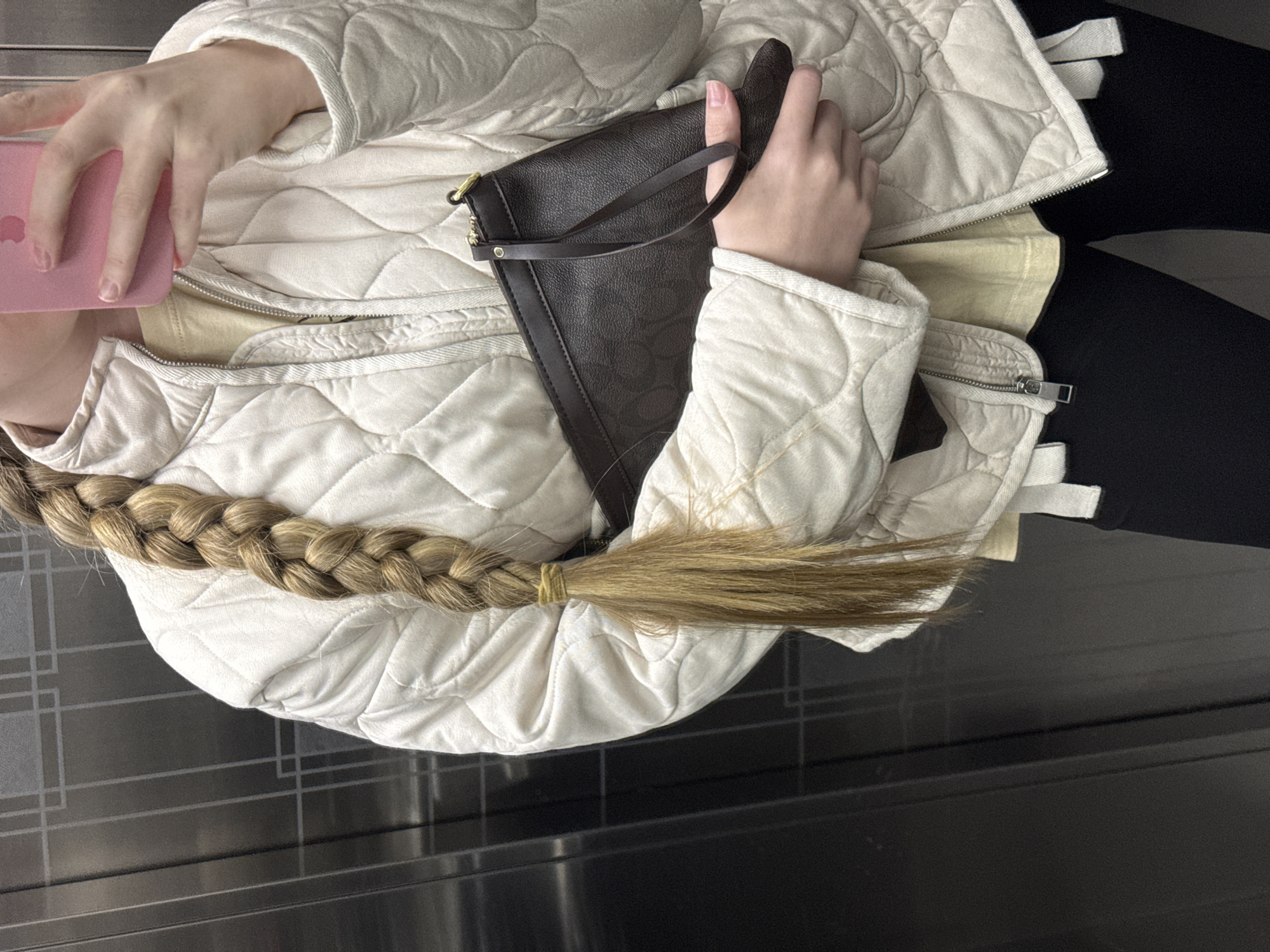 The image, taken from a high-angle perspective, captures a partially visible person in what appears to be an indoor setting in Petropavlovsk-Kamchatsky, Russia. The individual is wearing a light-colored, possibly cream or off-white, quilted jacket over a light yellow t-shirt, with dark leggings or trousers visible on the right.
The person holds a bright pink smartphone, identified as an iPhone by the visible Apple logo on its back, in their left hand. Their right hand clasps a small, dark brown handbag or clutch, which features a wrist strap and a subtle pattern.
A significant detail is the individual's long, light brown or blonde hair, styled into a thick braid that rests across the quilted jacket. The braid is secured near its end with a yellow hair tie, from which the rest of the hair cascades freely.
The background shows a dark floor with a geometric pattern of lighter lines, suggesting a modern, possibly public, interior space such as a waiting area or public transport. The scene depicts the person engaged in a casual activity, possibly using their phone or simply waiting. No specific time of day, weather, or emotions are discernible from the image.
