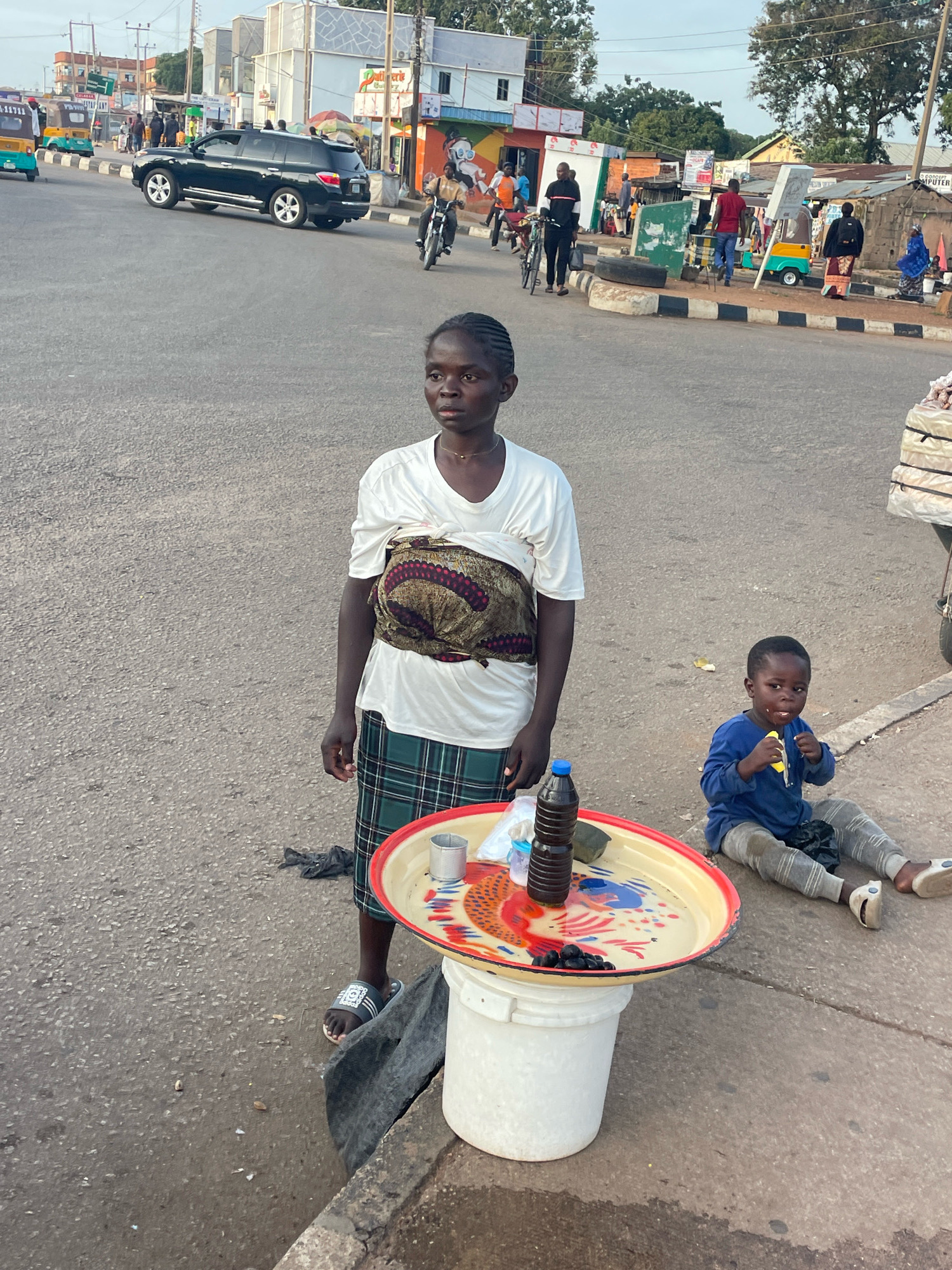A young woman and child are seen on a street in Barakin Lerawi, Nigeria. The woman stands in the foreground, wearing a white t-shirt, a patterned cloth wrapped around her waist, and a plaid skirt. She is looking towards the right of the frame. In front of her, a decorative tray sits atop a white bucket, holding a bottle of dark liquid, a metal cup, and some black berries. To the right of the woman, a young child in a blue long-sleeved shirt and striped pants sits on the ground, eating. In the background, a black SUV drives on the street, while various shops and people are visible on the sidewalk. A motorcycle is also present on the road. The environment appears to be a busy street with a mix of traffic and pedestrian activity.