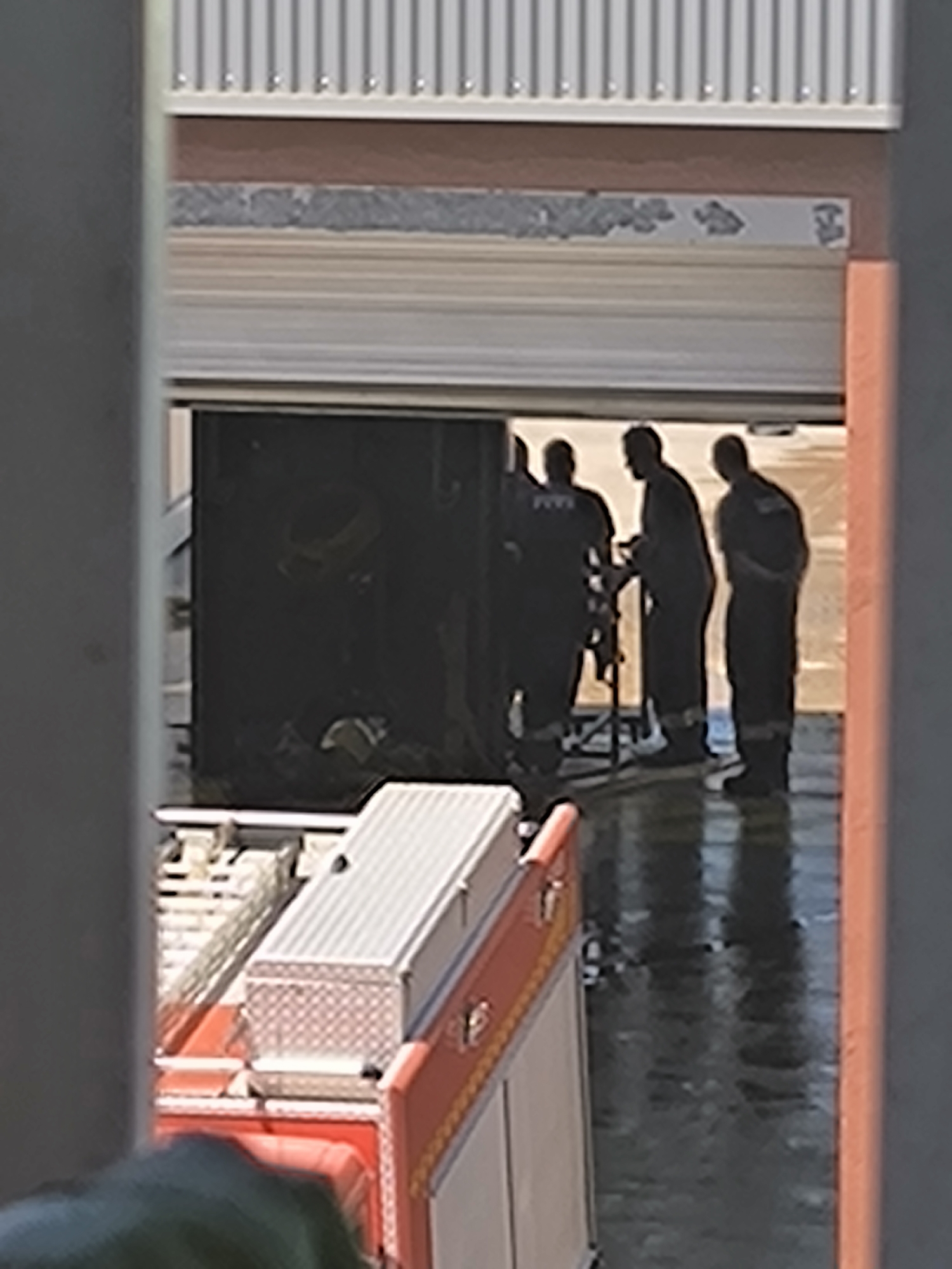 The image captures a scene within what appears to be an emergency services facility, likely a fire station, in Vilafranca del Penedès, Spain. Three male figures, silhouetted against a bright background, are gathered inside a large bay or garage opening. They are dressed in dark uniforms, possibly firefighter turnout gear or work coveralls, with one figure on the left showing a faint, light-colored patch or emblem on his back. They appear to be interacting with or inspecting a piece of equipment, possibly a portable stand or tool, positioned between the two central figures.
In the foreground, partially visible on the left, is the top section of a red and white emergency vehicle, consistent with a fire truck. It features a checkered pattern on a storage box or compartment. The floor of the bay is dark and highly reflective, suggesting it is wet or freshly cleaned.
Above the large opening, a multi-layered wall construction is visible. It includes a section of corrugated metal siding at the very top, followed by a brown horizontal band, a textured grey band, and then a white horizontal strip directly above the garage door. On this white strip, there are faint, partially obscured, and possibly faded or damaged dark markings. While indistinct, these markings appear to be text or a logo. On the left side of the strip, some fragmented characters are visible, potentially part of a word like "GOMAR" or similar. A smaller, blurry graphic or logo is present on the right side of the strip.
The bright lighting suggests it is daytime, casting the figures into silhouette. No specific emotions are discernible. The overall setting implies a period of readiness or maintenance within an emergency response context.