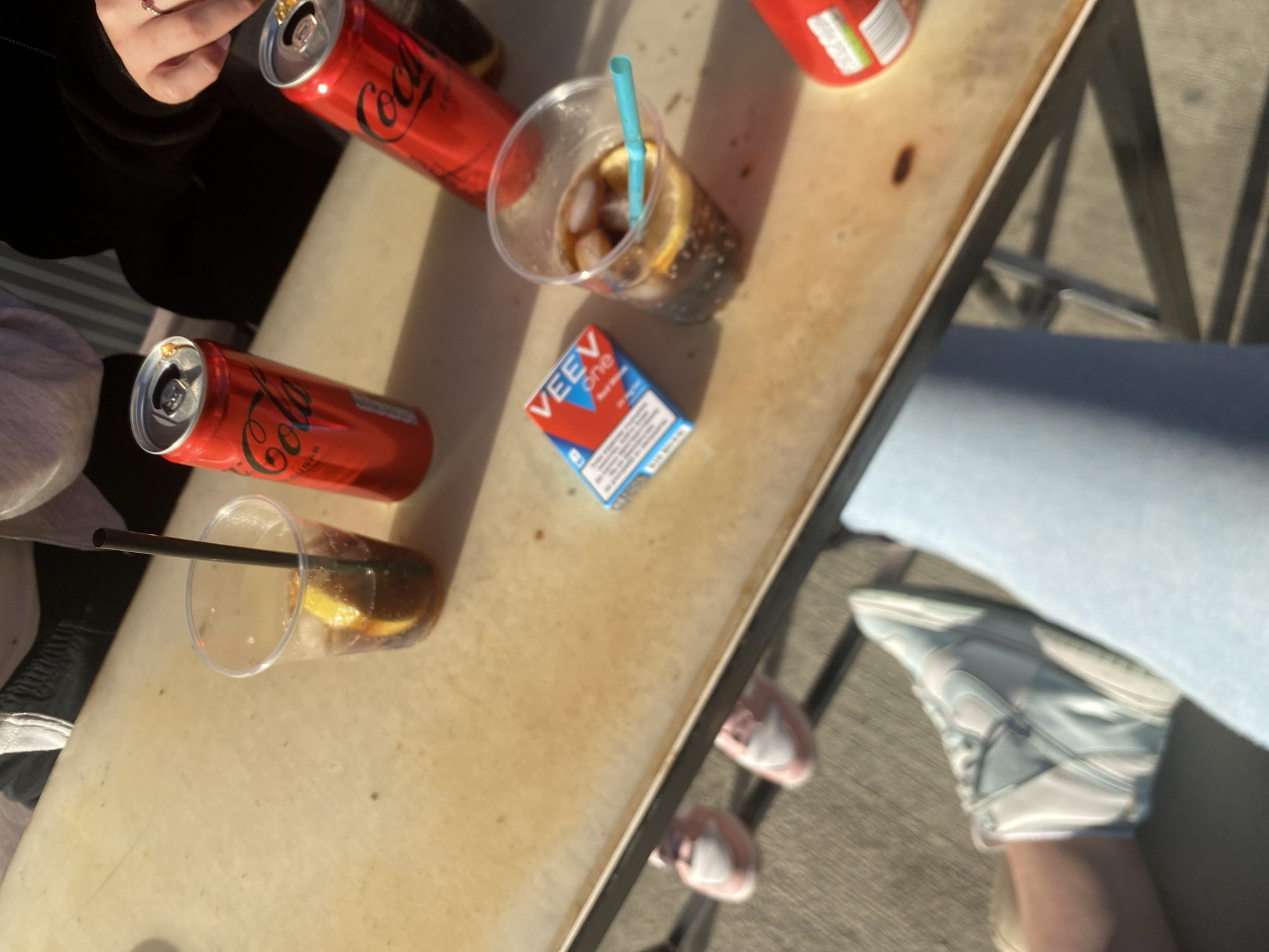 On a light-colored table, possibly in Satovcha, Bulgaria, sit two open cans of Coca-Cola, two clear plastic cups filled with a dark liquid and lemon slices, and a pack of VEev One e-cigarettes. Two individuals are present, indicated by a hand wearing a ring, a partial view of clothing, and feet wearing sneakers. The environment appears to be outdoors with sunlight and shadows visible.