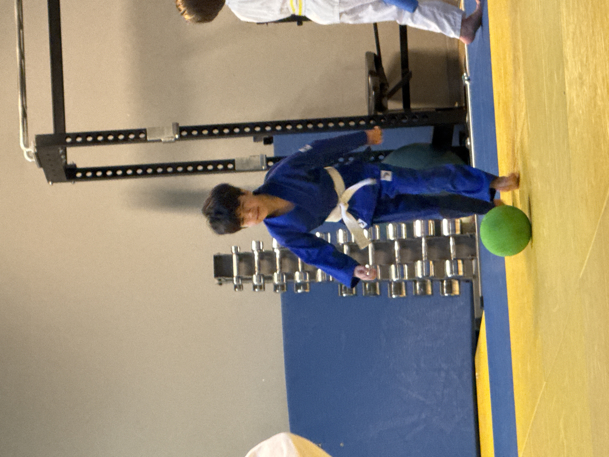 A young boy, wearing a blue judo gi with a white belt, is playing with a green ball on a yellow mat. He is looking towards the ball, with his right foot extended forward as if to kick it. In the background, a rack of weights and a blue wall are visible. To the right, another child in a white judo gi with a yellow belt is partially visible. The scene appears to be a gymnasium or martial arts training facility. The location is not specified in the image or its description.