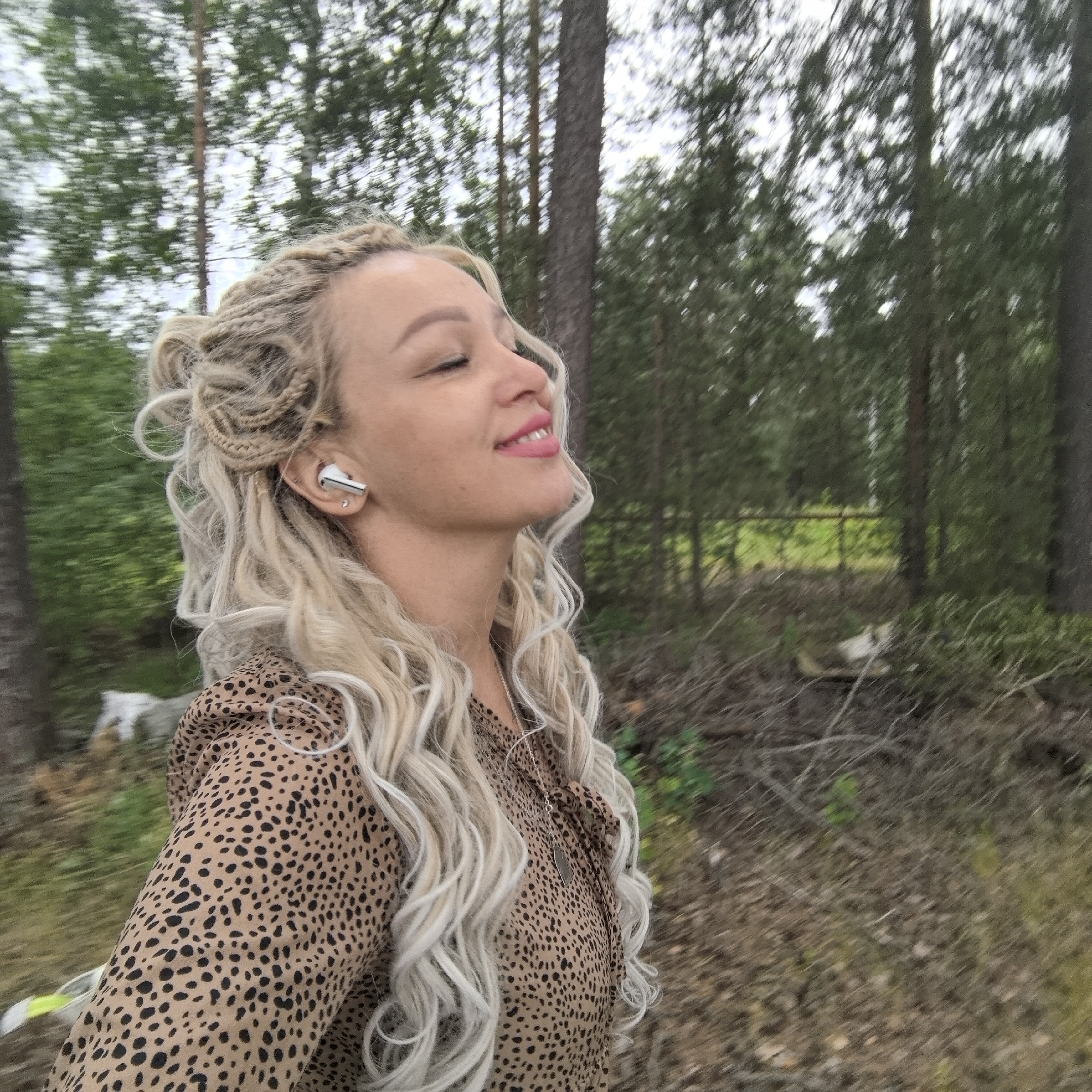 In Noviy, Russia, a young woman with blonde hair styled in braids and loose curls is captured in a side profile outdoors, amidst a forest setting. She is wearing a tan and black spotted top and a silver necklace. A white earbud is visible in her left ear. Her eyes are closed, and her head is tilted upwards, suggesting a moment of enjoyment or relaxation. The background is filled with trees, with a hint of a fence and greenery visible further back. The ground appears to be covered in dry leaves and twigs. The lighting suggests an overcast day.