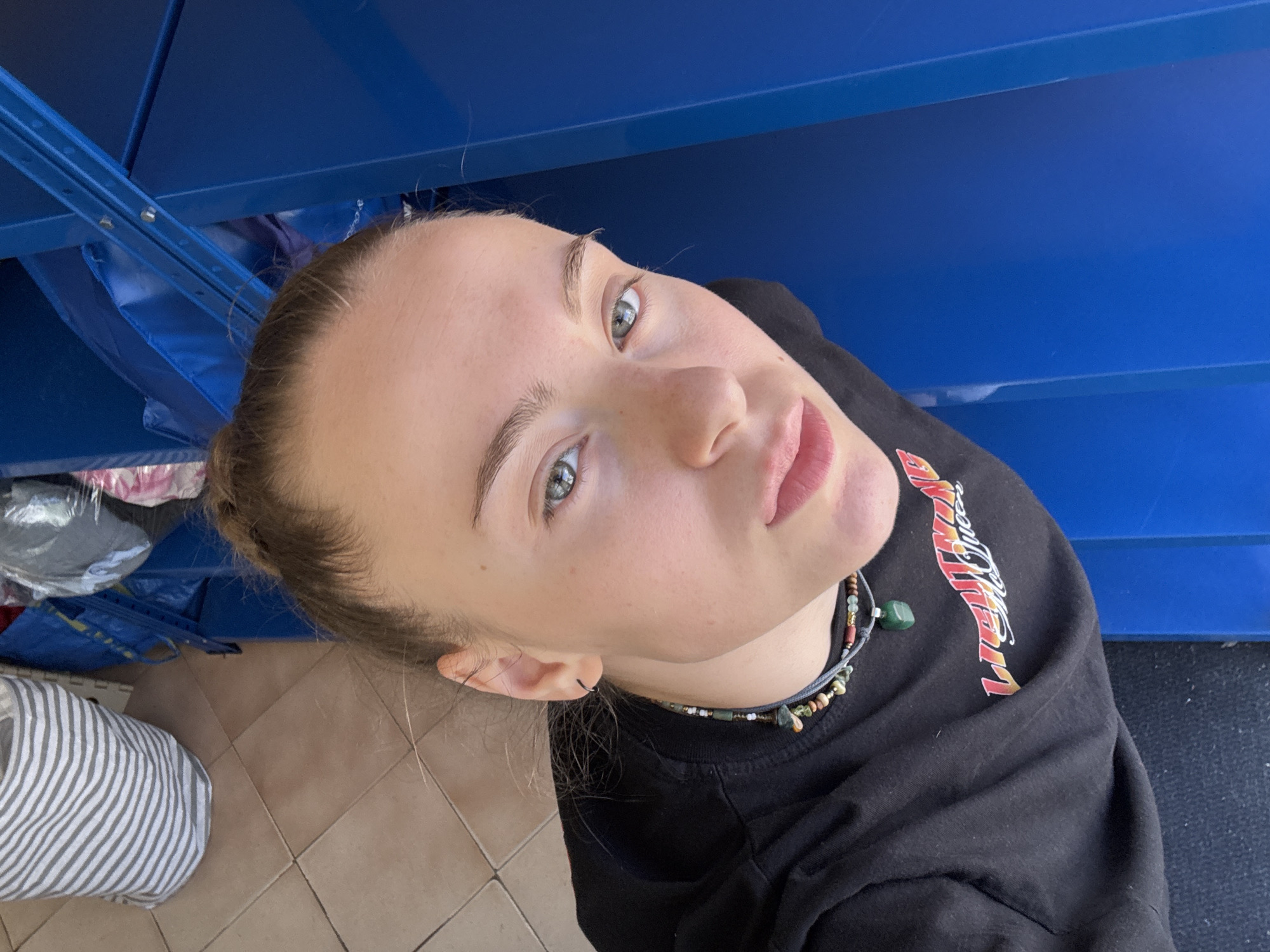 The image, taken from a high-angle, selfie-style perspective, features a young woman with her hair pulled back in a bun. She is looking directly at the camera, her expression neutral. She is wearing a black t-shirt with a graphic print on it and a beaded necklace. The background shows a tiled floor, a striped laundry bag, blue plastic shelving, and some clothing items. The scene is indoors, with natural light illuminating the subject. There is no indication of the specific location within the city of Sofia, Bulgaria, or any forensic evidence.