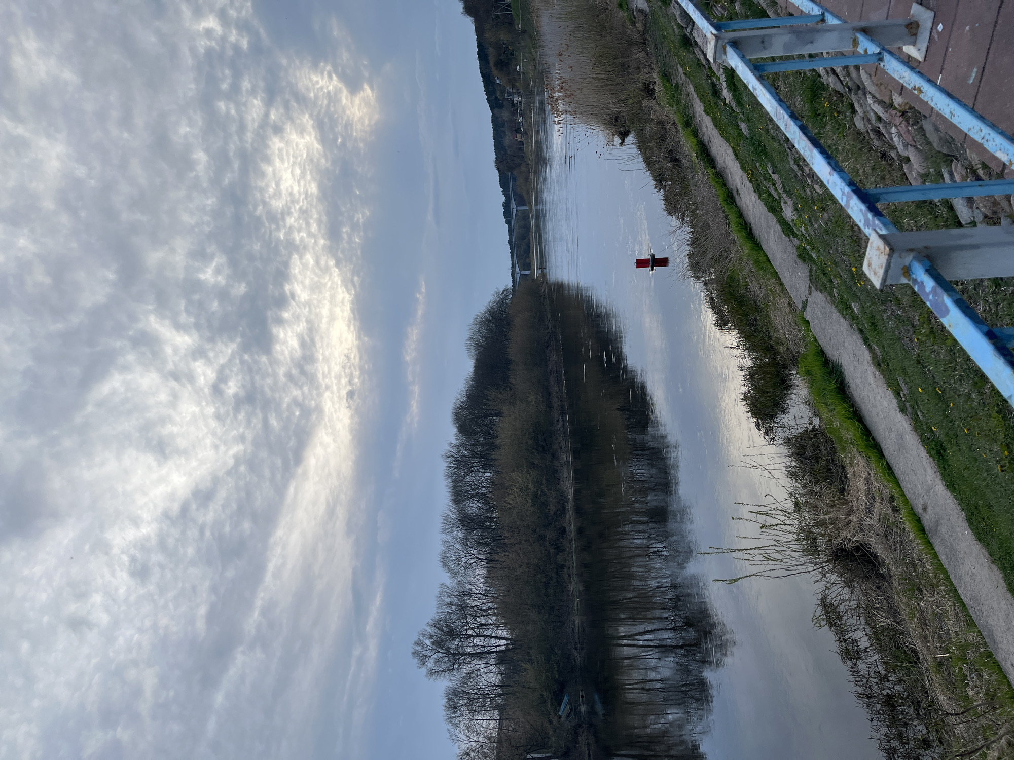 The image shows a canal with trees reflected in the water. A red buoy is floating in the canal. A blue metal railing borders the canal path, which has grass and concrete segments. The sky is cloudy with some bright patches. A bridge is visible in the distance. The location and city are unknown.