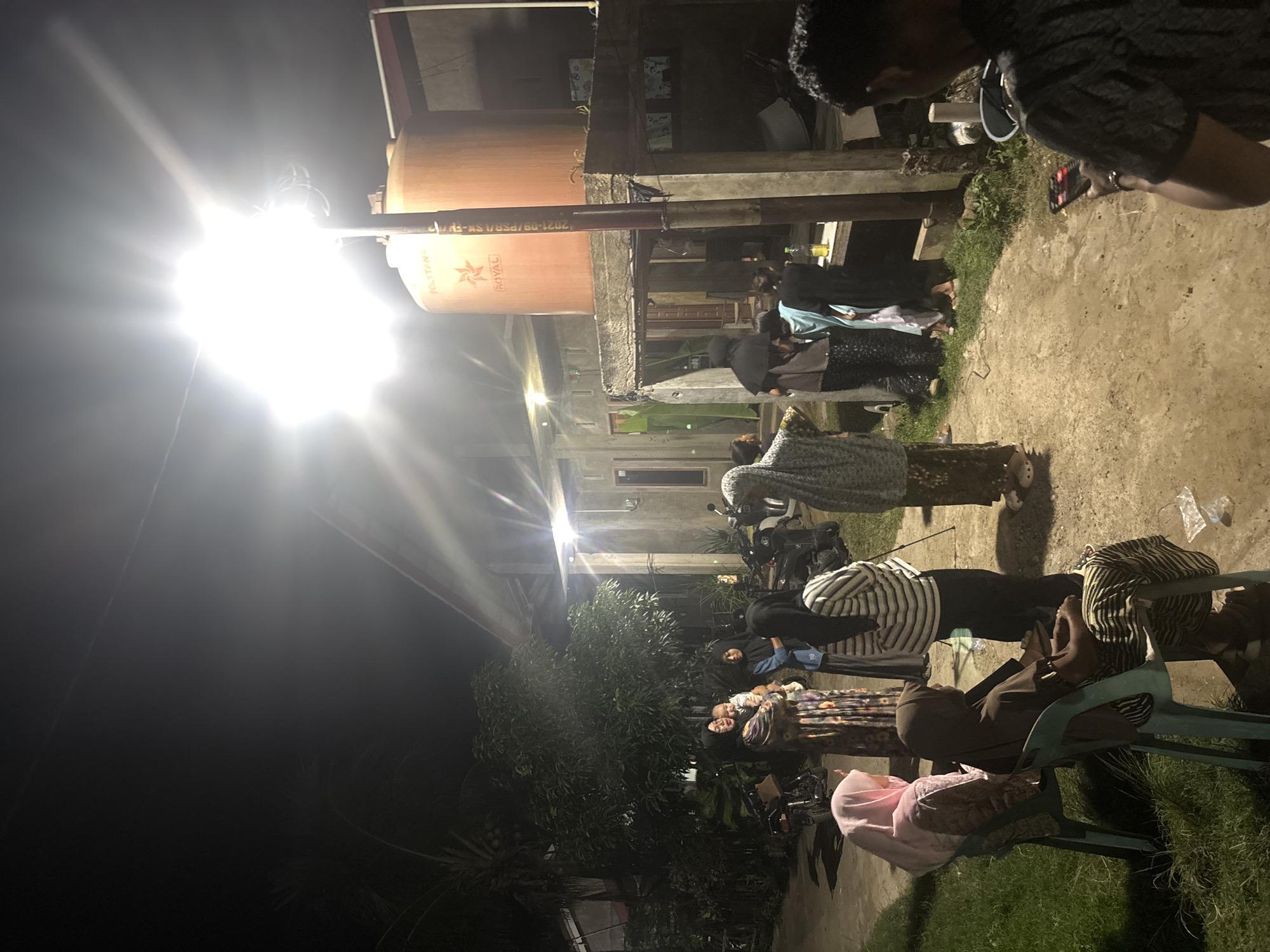 A group of people, mostly women and children, are gathered outdoors at night in what appears to be a residential area in Lhokseumawe, Indonesia. A bright overhead light illuminates the scene. Several individuals are wearing headscarves and traditional clothing. Some are standing and conversing, while others are seated on plastic chairs. A large orange water tank is visible in the background, mounted on a concrete structure. Motorcycles are parked nearby. The ground is a mix of dirt and grass. One person in the foreground is holding a smartphone. The overall atmosphere suggests a casual social gathering.