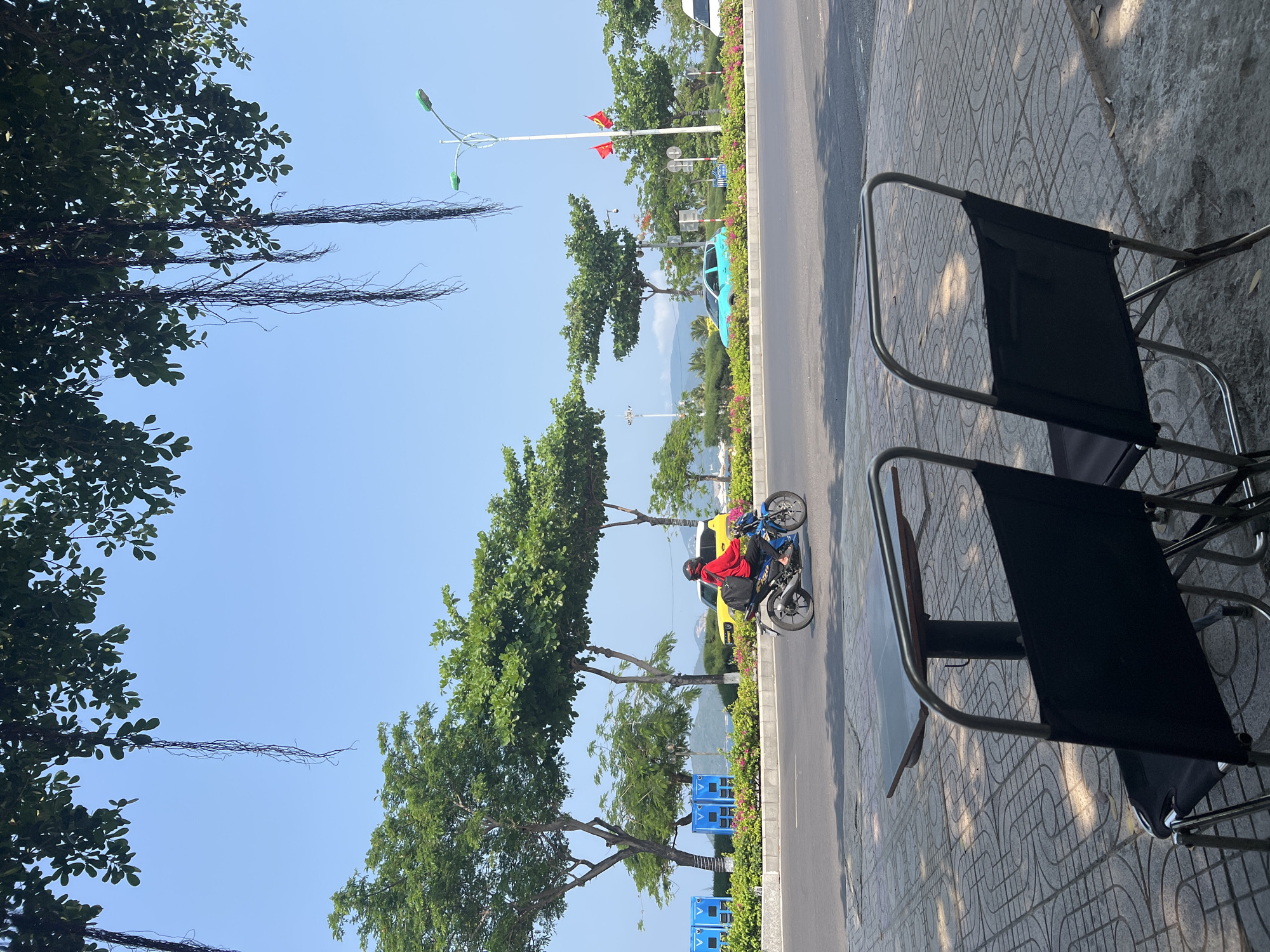 The image captures a street scene in Nha Trang, Vietnam, viewed from a partially shaded outdoor cafe. Two black folding chairs with silver metal frames are in the foreground, sitting on a patterned concrete sidewalk. A street runs parallel, lined with flowering bushes and small trees. A person wearing a red shirt and helmet is riding a blue motorbike along the road. Further down the street, a yellow vehicle is followed by a turquoise vehicle. A tall, white lamppost with green lamps and Vietnamese flags can also be seen. The sky is blue and the scene is bathed in bright sunlight. A large tree with dangling aerial roots partially obstructs the view on the left side of the frame.