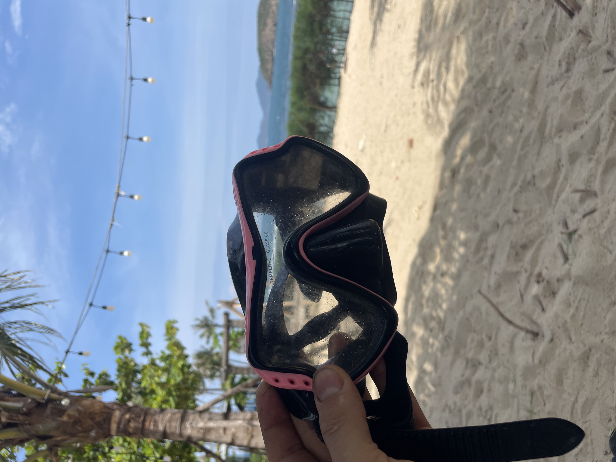The image shows a person holding a pink and black snorkeling mask in their right hand against a backdrop of a beach scene, likely in Bac Nha Trang Ward, Vietnam. The mask is slightly tilted, showing reflections in the lens. The background includes a sandy beach, turquoise water, a distant mountain range, trees, and a string of lights hanging overhead. The hand holding the mask is partially visible, and the snorkel is attached to the mask.