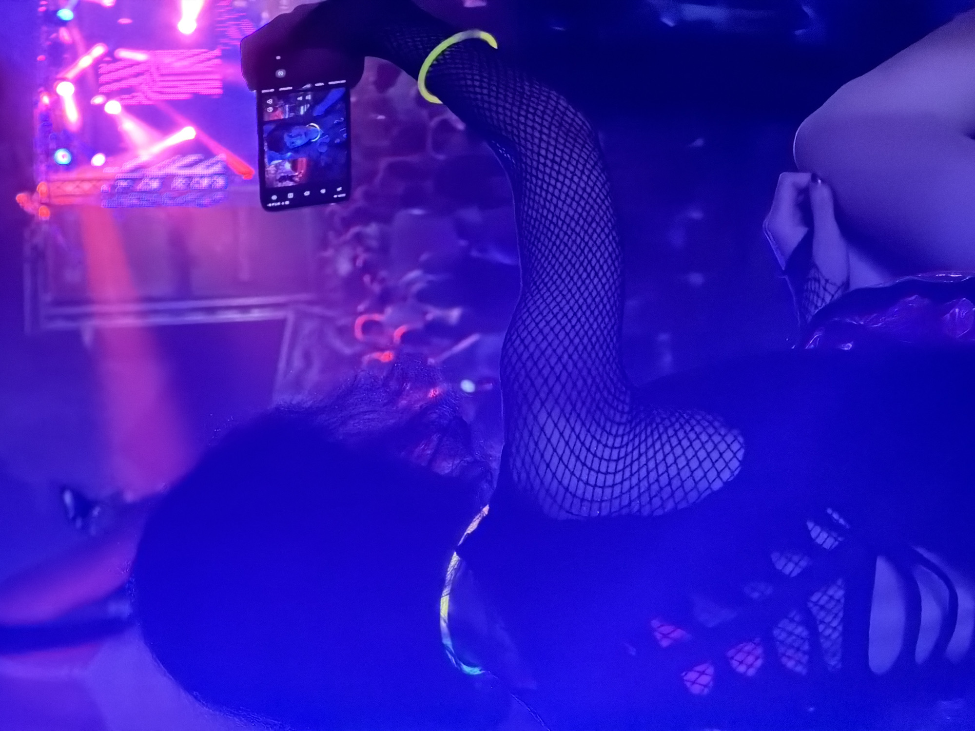The image depicts a person holding a smartphone in their right hand, taking a selfie at what appears to be a concert or club. The person is wearing a black, ripped fishnet top, and neon glow bracelets around their wrist and neck, indicating a party or festival atmosphere. The background is filled with a crowd of people and stage lighting, characterized by vibrant pink and blue hues, suggesting a low-light, energetic environment. The smartphone screen shows a selfie of the person, with a faint glimpse of another individual in the background. The overall scene is dimly lit, with strong artificial lighting creating a dramatic and colorful ambiance. The location is specified as the city of Echternach, Luxembourg, but no specific landmarks or identifying features of the city are visible in the image.