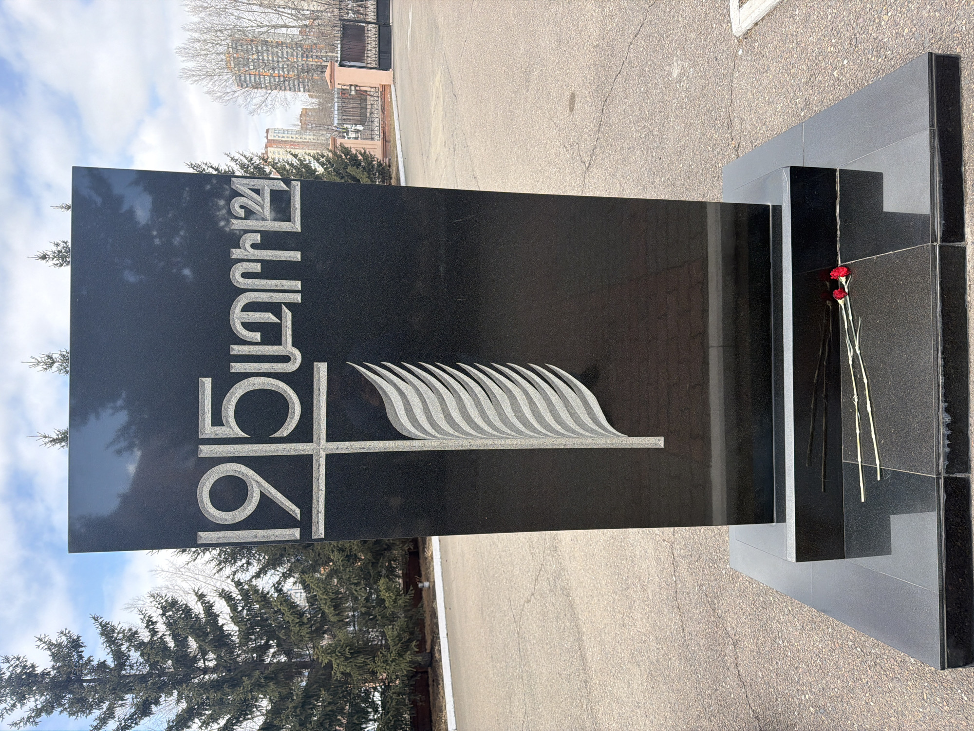 A tall, black, polished stone monument stands prominently in an outdoor setting in Krasnoyarsk, Russia. The monument, likely made of granite or similar material, features deeply carved, light-colored text and an emblem. The text, in Armenian script, reads "1915 ՑԵՂԱՍՊԱՆՈՒԹՅՈՒՆ," which translates to "1915 Genocide." Below the numbers and letters, an emblem is carved, depicting a stylized cross combined with a flowing, multi-layered design resembling flames or a rippling flag.
At the base of the monument, on a lower, multi-tiered platform of similar dark stone, two red carnations are gently placed, their stems reflecting on the polished surface. The ground around the monument is paved with asphalt, showing some cracks. In the background, there are several trees; some are evergreen pines, while others are deciduous and bare, suggesting late winter or early spring. Beyond the trees, various buildings are visible, including one with a reddish-brown facade and another taller, more modern structure. A dark metal fence and gate are also part of the distant scene. The sky above is partly cloudy with patches of blue visible. The overall scene evokes a solemn atmosphere of remembrance and commemoration.