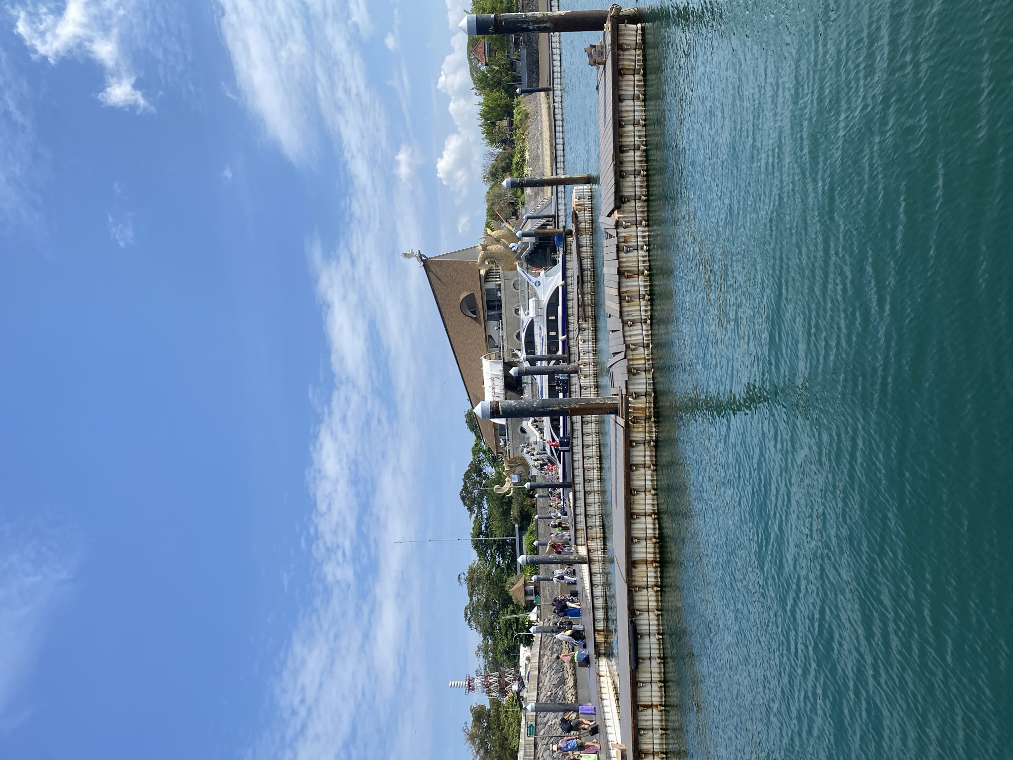 A ferry terminal in Denpasar, Indonesia, is shown on a bright, sunny day with a clear blue sky filled with scattered white clouds. In the foreground, turquoise water laps against a wooden dock where several people are disembarking from a white ferry. The people are dressed in casual clothing and are pulling luggage. The dock extends across the width of the image, supported by metal pylons. In the background, a large, modern building with a distinctive roof design featuring golden dragon sculptures and a bird ornament is visible. To the left of the building, a tall, red and white striped tower with a spiral structure stands against the sky. Lush green trees line the background, creating a tropical atmosphere.