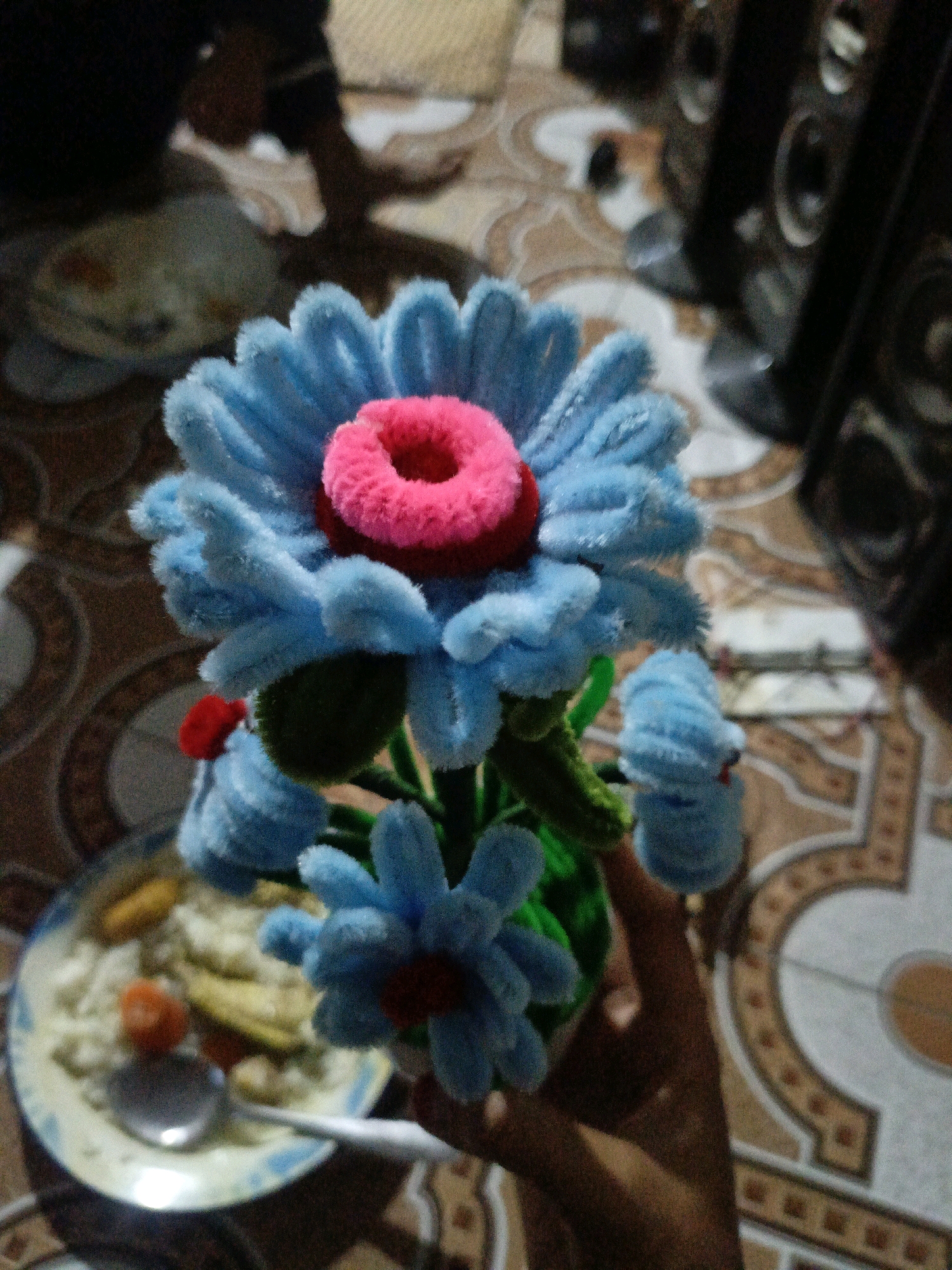 An overhead, slightly blurry, indoor, low-light shot depicts a hand holding a small, artificial blue flower with a pink center, made from what appears to be pipe cleaners. The flower is held in front of a plate of food, which includes rice, what look like slices of banana, and possibly some small pieces of carrot or other vegetables. The background shows a patterned tiled floor and what appears to be a large speaker system. The scene suggests a domestic setting, possibly in Pagar Ayu, Indonesia, where someone is presenting or showing the artificial flower alongside a meal.