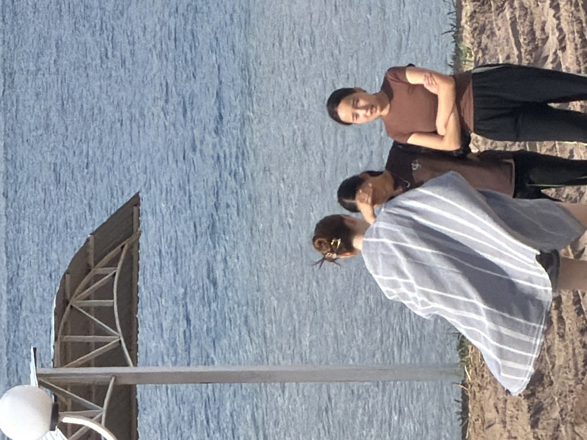 A group of three young women are standing on a sandy area near a body of water. One woman in the foreground is seen from behind, wearing a light blue and white striped garment. To her right, another woman, wearing a brown t-shirt and black pants, has her arms crossed and is looking towards the camera. Behind her, a third woman, wearing a brown t-shirt, has her hand covering her face. A lamppost with a white globe light and a metal canopy stands to the left of the frame. The background is dominated by a large expanse of blue water. The lighting suggests it is daytime.