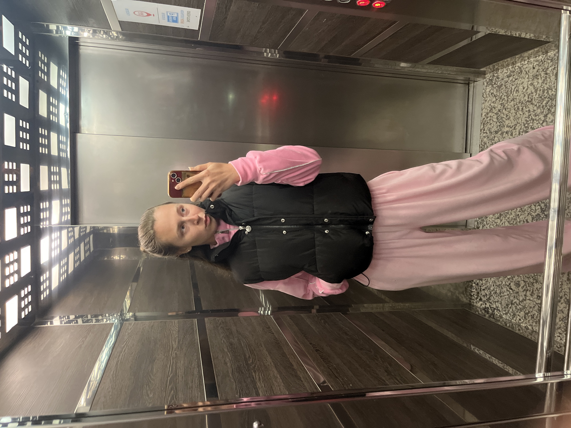 A young woman is taking a selfie in an elevator. She is wearing a pink tracksuit and a black vest. The elevator has mirrored walls and a wood-paneled ceiling. The location is Rafailovići, Montenegro. The elevator appears to be in a building, based on the lights in the elevator shaft.