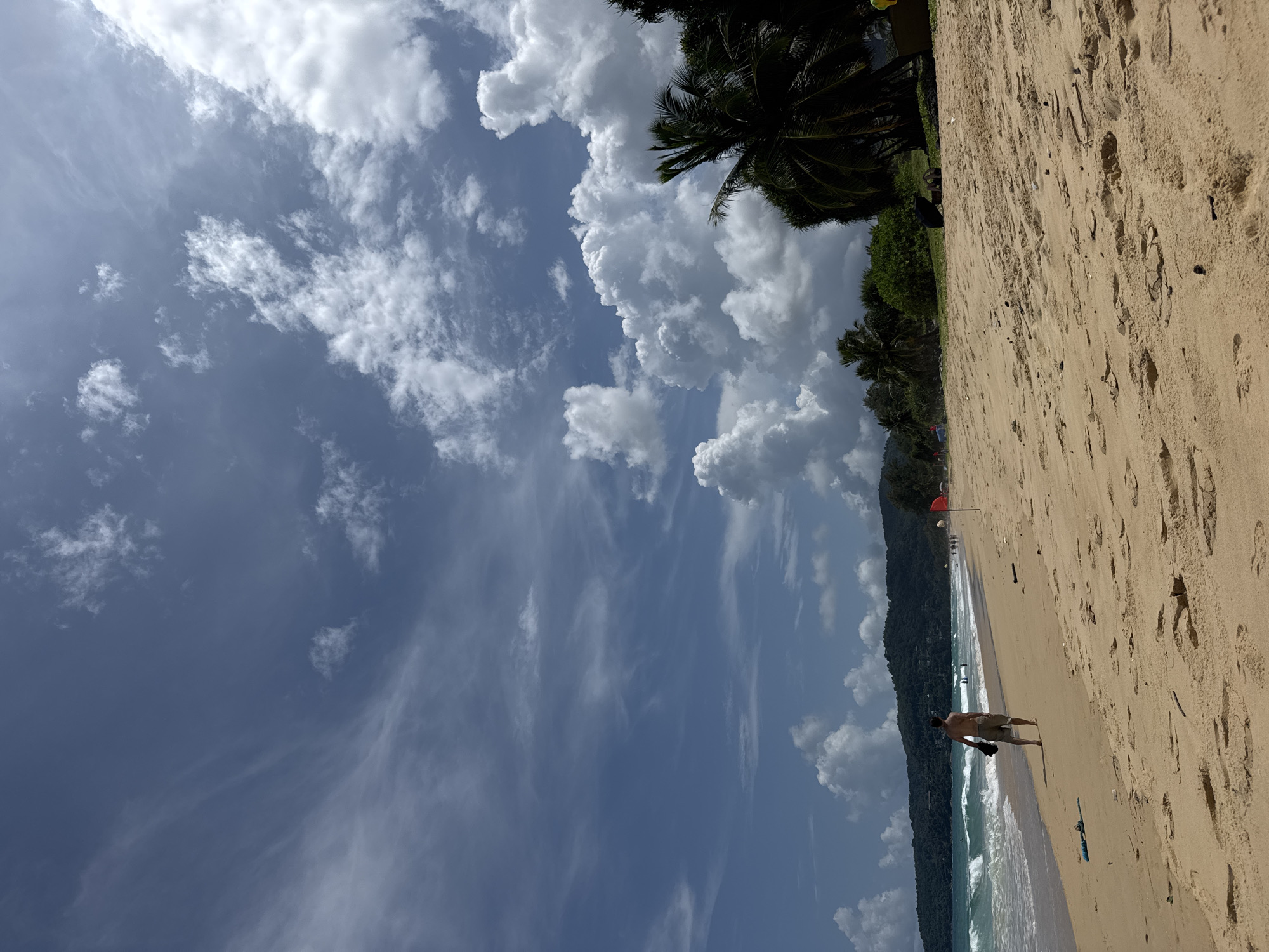 A man walks along a sandy beach in Karon, Thailand. The man is in the foreground and is wearing shorts and no shirt. He is walking away from the camera and towards the water. The beach is wide and sandy with a few scattered pieces of driftwood. In the background, there are waves crashing on the shore and a line of palm trees. The sky is blue with a few white clouds. There are other people walking along the beach in the distance and a red flag is planted in the sand. The overall scene depicts a peaceful day at the beach.