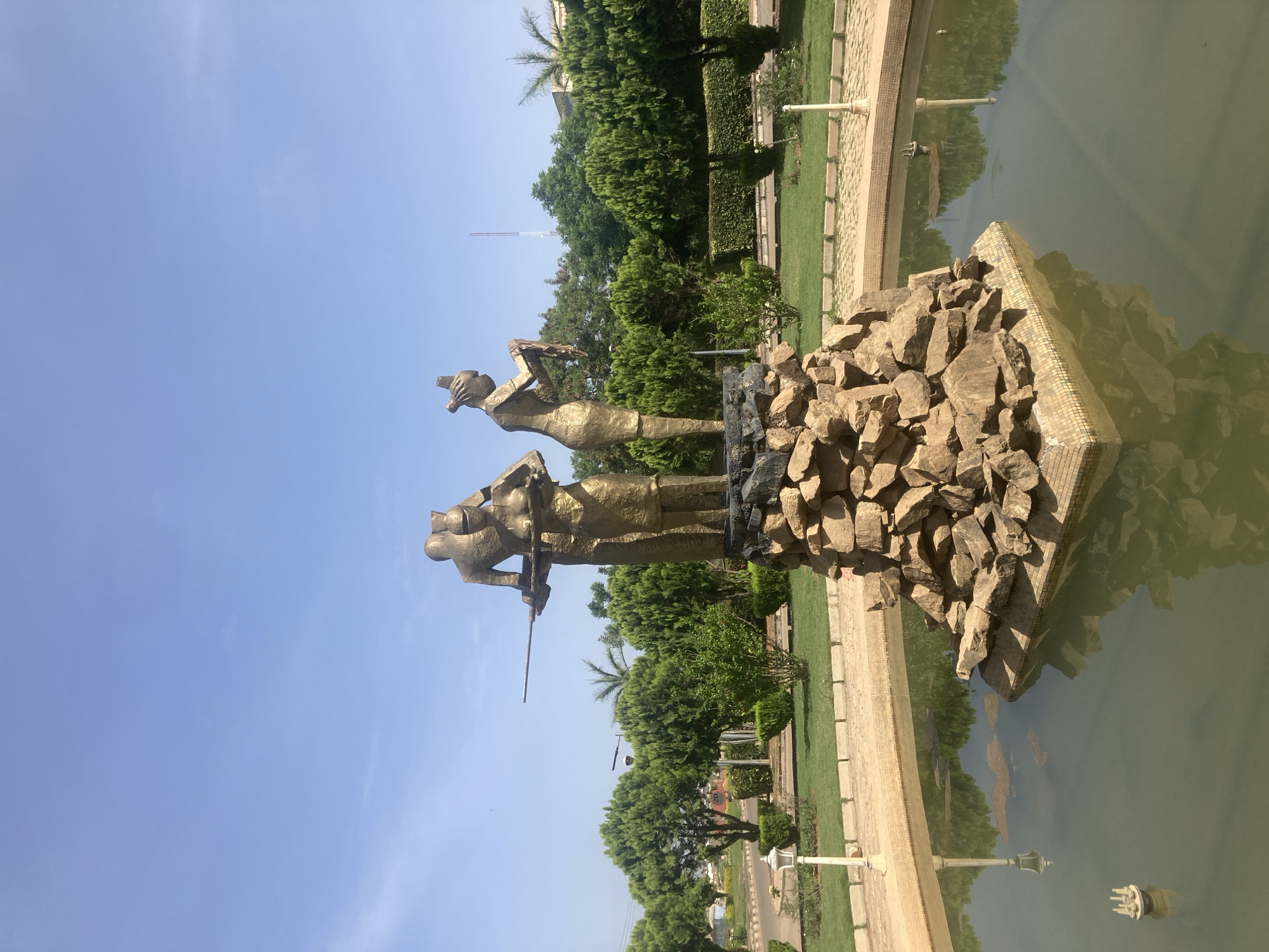 A vertical, eye-level outdoor shot of a bronze-colored statue of two men atop a rock formation, partially submerged in a rectangular body of water. The background features green trees and a clear blue sky. The statue depicts two men, one holding a device in his hands, standing above the rocks. The rock formation extends into the water, creating a reflection. The scene is situated in a park setting, with landscaped grass and light poles visible. The city of Gwong, Nigeria, is the probable location.