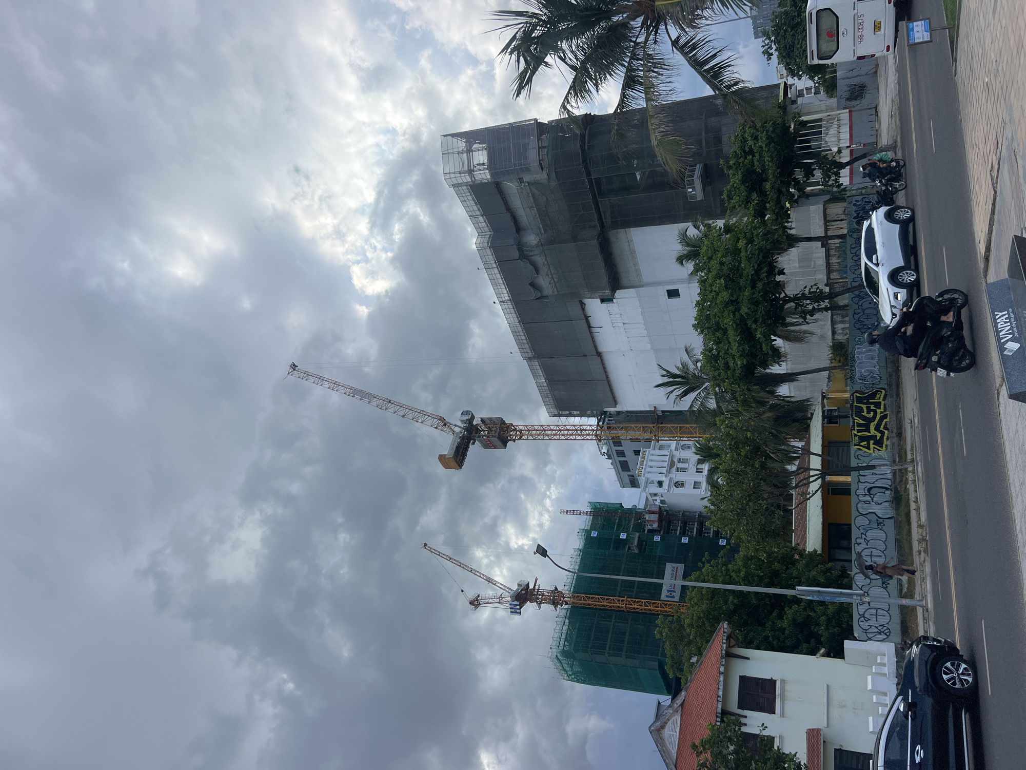 In an overcast urban scene, construction cranes tower over unfinished buildings. A tall, yellow crane on the left, with a white cabin, stands in front of a building covered in green netting. To its right, another, larger yellow crane with a yellow cabin is positioned in front of a multi-story structure wrapped in dark netting. In the foreground, a white SUV is parked, and a motorcycle passes by. A white bus with license plate "43B-008.15" is visible further down the road. A person walks with an umbrella on the sidewalk, which is adorned with graffiti. Palm trees and other foliage dot the landscape, with a white house in the lower left corner. The sky is filled with dramatic, dark clouds, suggesting impending or recent rain.