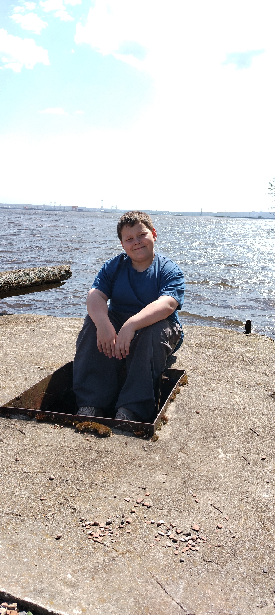 A boy sits in a square, metal-lined hole in a concrete pier or structure near water. The water is choppy and reflects sunlight. Industrial structures are visible in the distance across the water. The concrete surrounding the hole is cracked and has small pieces of material scattered on it. Moss is growing in the hole. The boy wears a blue t-shirt and gray pants. Based on industrial structures across the water, it appears this image was captured in the city of Noviy, Russia.