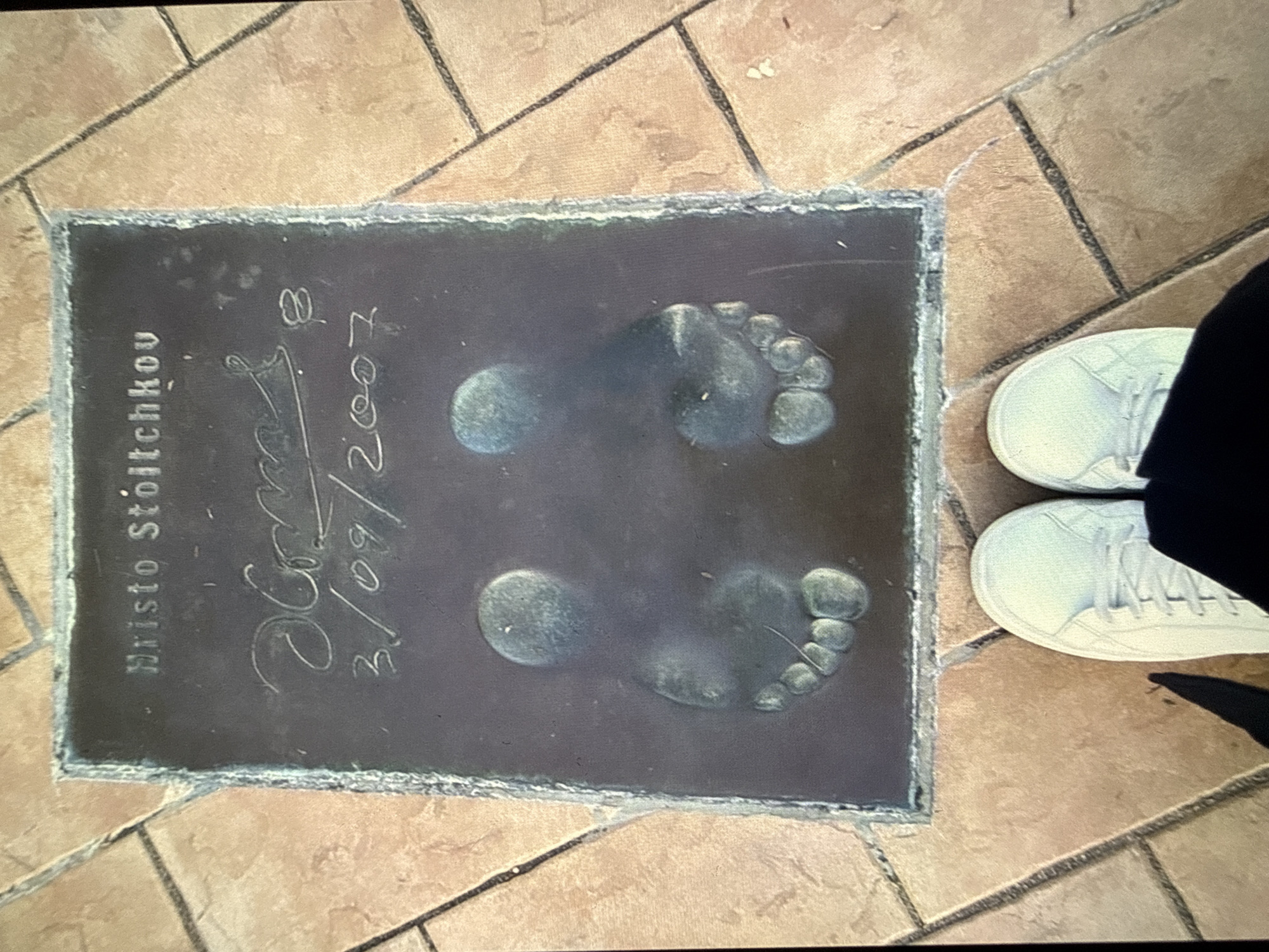 The image depicts a black slab set into a paved area, likely a public walkway, possibly in Sofia, Bulgaria. Inscribed on the slab are the words "Hristo Stoitchkov" above a signature and the date "3/09/2007." Imprints of two bare feet are visible on the slab. Adjacent to the slab on the paved walkway, a pair of white sneakers with laces are visible, positioned as if someone is standing next to the slab. The paving surrounding the slab consists of medium-sized square tiles with darker grout lines. The overall lighting suggests daylight.