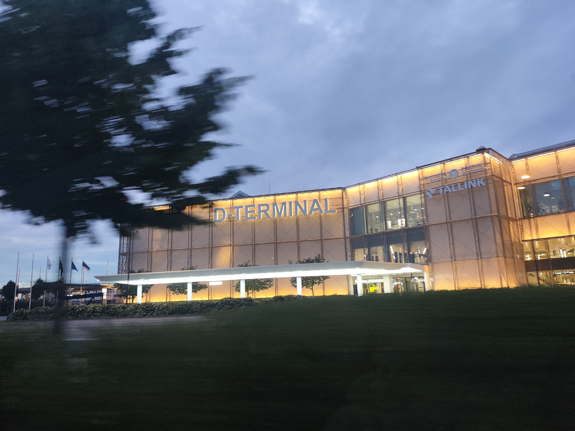The image shows a large, modern building with "D-TERMINAL" prominently displayed in large blue letters on its facade. Above the "D-TERMINAL" sign, the text "TALLINK" is also visible. The building appears to be a ferry or cruise terminal, likely in Tallinn, Estonia, given the presence of Estonian flags on flagpoles in the background. The architecture is characterized by large glass windows and a facade covered in a light-colored material, possibly some type of fabric or screening, with warm yellow lights illuminating the exterior.
In front of the building, there is a covered walkway or entrance area, supported by white pillars, with several trees planted along its edge. Beyond this, a manicured lawn or grassy area stretches towards the foreground. A blurred image of a tree or foliage is visible in the left foreground, indicating that the photo was taken from a moving vehicle.
The sky is overcast with dark clouds, suggesting it is either early morning or late evening, likely during dusk. The overall atmosphere is somewhat somber due to the lighting and weather conditions, but the illuminated building provides a sense of warmth and activity. No people are clearly visible in the image, and the focus is on the architecture and signage of the terminal.