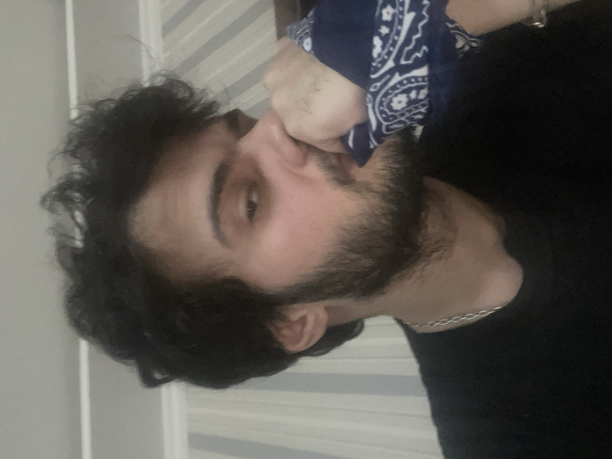 A medium shot captures a young man with dark, curly hair and a beard, looking towards the camera with his right hand covering his mouth and nose with a blue and white paisley bandana. He is wearing a black t-shirt and a silver chain necklace. The background consists of a wall with vertical striped wallpaper and a white molding. There are no visible indications of the city of Tbilisi, Georgia, in the image.