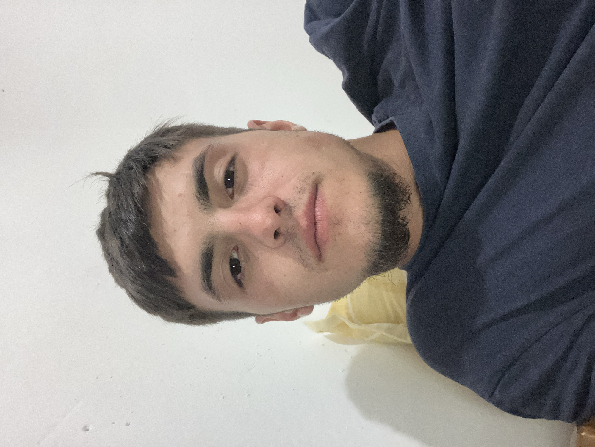 A medium close-up shot of a man's face and upper torso, rotated 90 degrees counter-clockwise. The man has short, dark brown hair and a short, scruffy beard. He is wearing a navy blue t-shirt. The background is a white wall with some faint imperfections. A hint of a light yellow object is visible behind his shoulder. Location: Unknown, including if it is located in the city of Komuniga, Bulgaria.