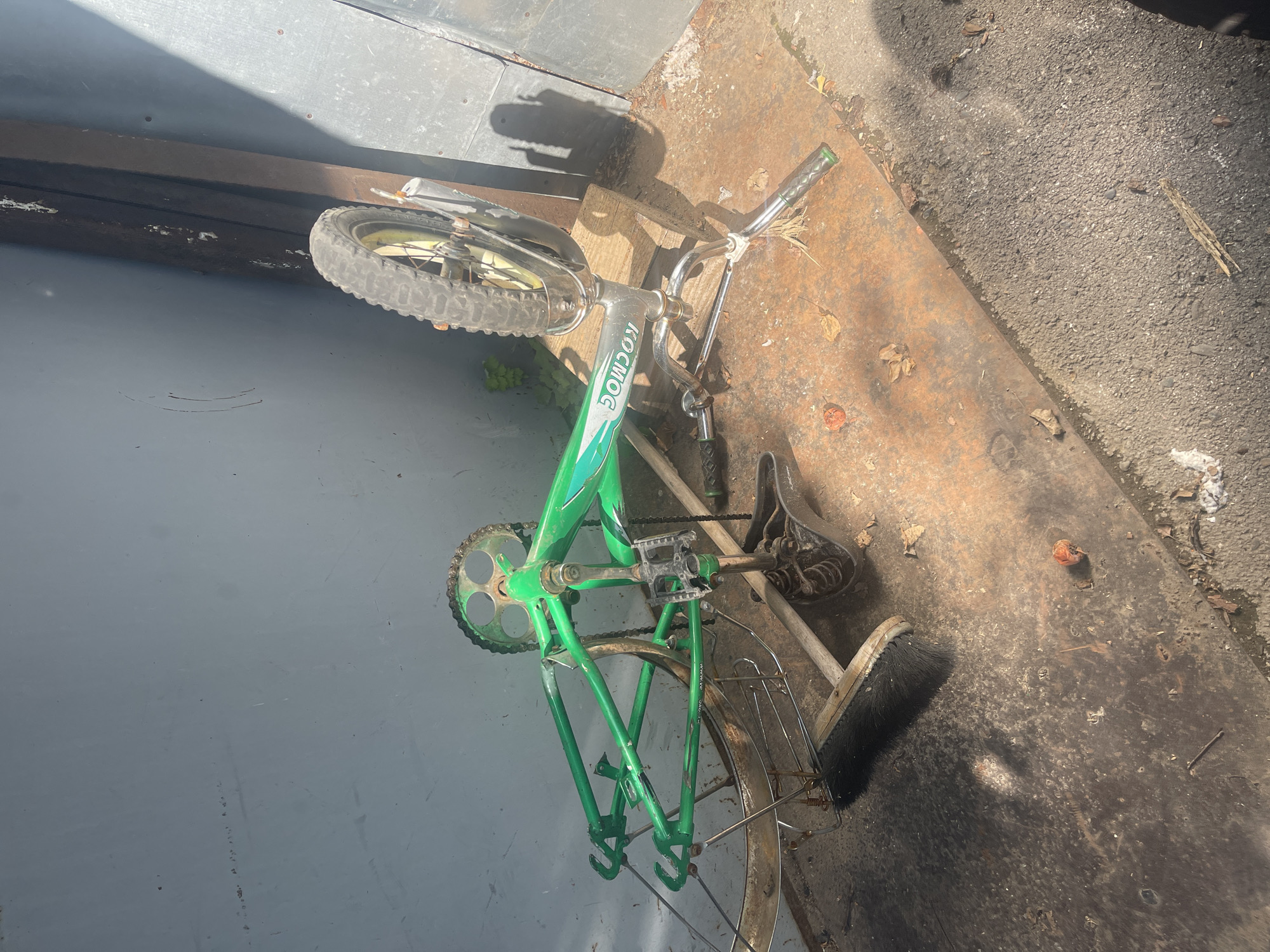 The image depicts a dilapidated green bicycle lying on its side on a rusty metal surface. The bicycle is missing its front wheel and appears to be in a state of disrepair. The background shows a grey, textured wall and a vertical metal beam. The ground is covered with debris, including leaves and dirt, suggesting an outdoor or neglected environment. Sunlight casts shadows on the scene. The bicycle's frame is visible, along with its pedals, chain, and a single rear wheel. The word "KOSMOC" is visible on the frame. The scene appears to be in the city of Sibirskiy, Russia, based on the provided context.
