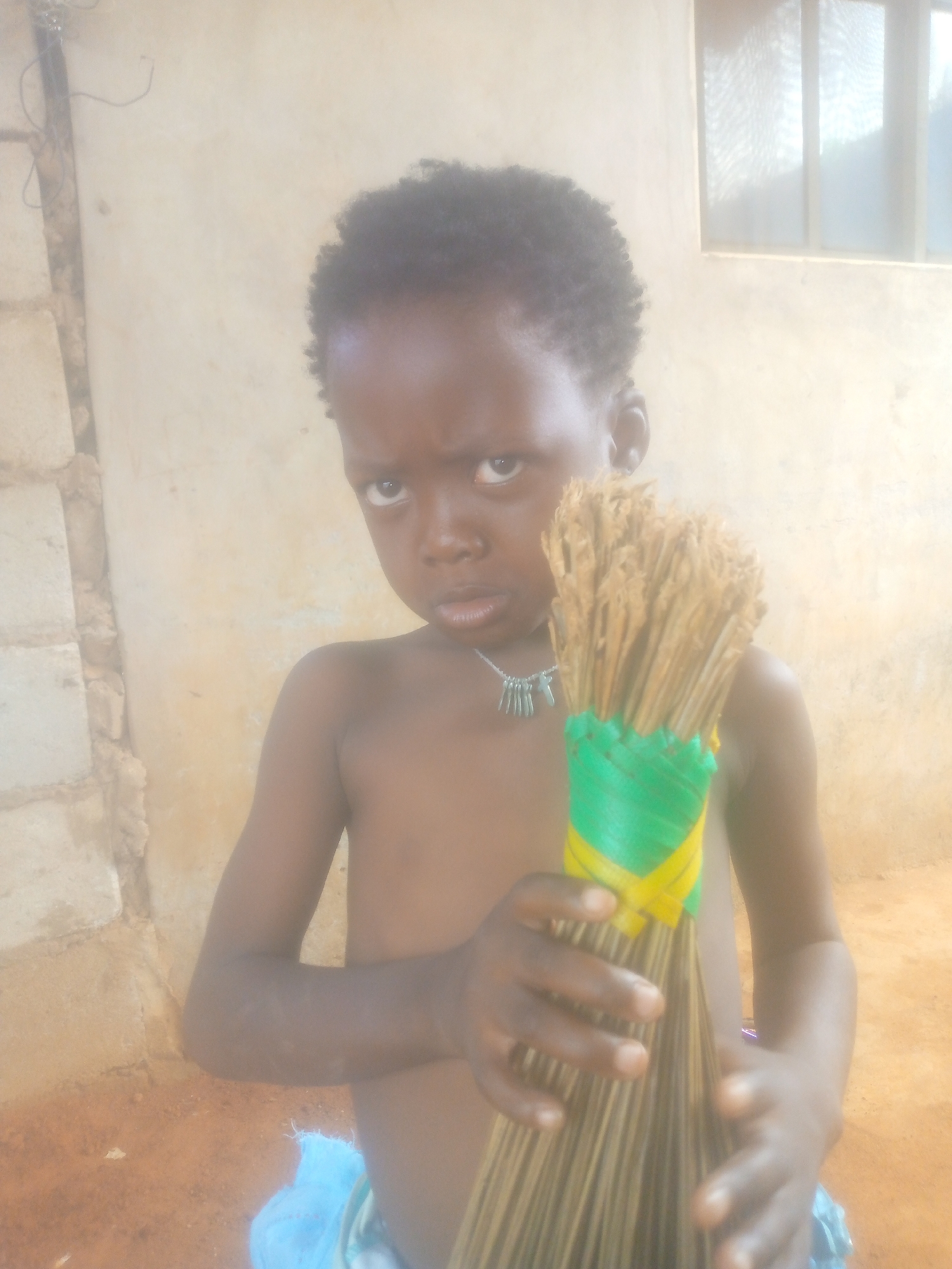 A young child, appearing to be between 4 and 7 years old, stands looking directly at the camera with a serious and slightly intense expression. The child has dark skin and short, curly black hair. They are shirtless, wearing a light-colored garment around their waist, and a necklace adorned with several small, metallic, cylindrical charms.
The child is holding a handmade whisk or small broom, fashioned from bundles of dried, slender natural fibers. The handle of the whisk is distinctively wrapped with bright green and yellow woven or plastic strips.
The setting appears to be a simple, perhaps indoor or semi-enclosed space. The background features a light-colored wall, possibly rendered or painted. To the left, a section of a concrete block wall is visible, hinting at the structure's construction. In the upper right corner, a window with multiple panes allows bright daylight to stream in, suggesting it is daytime. The ground visible at the bottom of the frame is reddish-brown earth. The overall scene suggests a candid portrait captured in a modest, possibly rural, environment.
No visible text is present in the image.
Location context: Unknown, Unknown