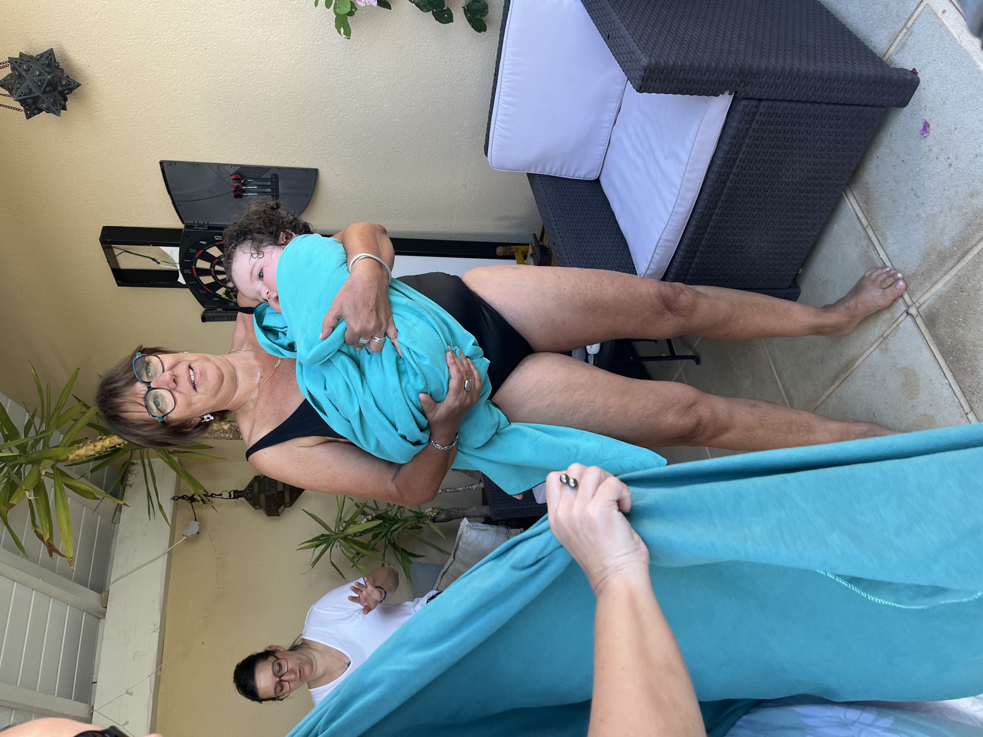 In an outdoor patio setting, likely in Saint-Galmier, France, a woman with glasses and a black swimsuit holds a baby wrapped in a turquoise blanket. The baby has curly dark hair and is looking to the side. Another woman, also wearing glasses and a white t-shirt, stands in the background to the left, gesturing with her hands. To the right, a dark wicker chair with white cushions is visible, and a dartboard hangs on the wall. A third person, whose hands are visible in the foreground, is holding up a large piece of turquoise fabric. The environment suggests a warm, possibly sunny day, with tiled flooring and some potted plants.