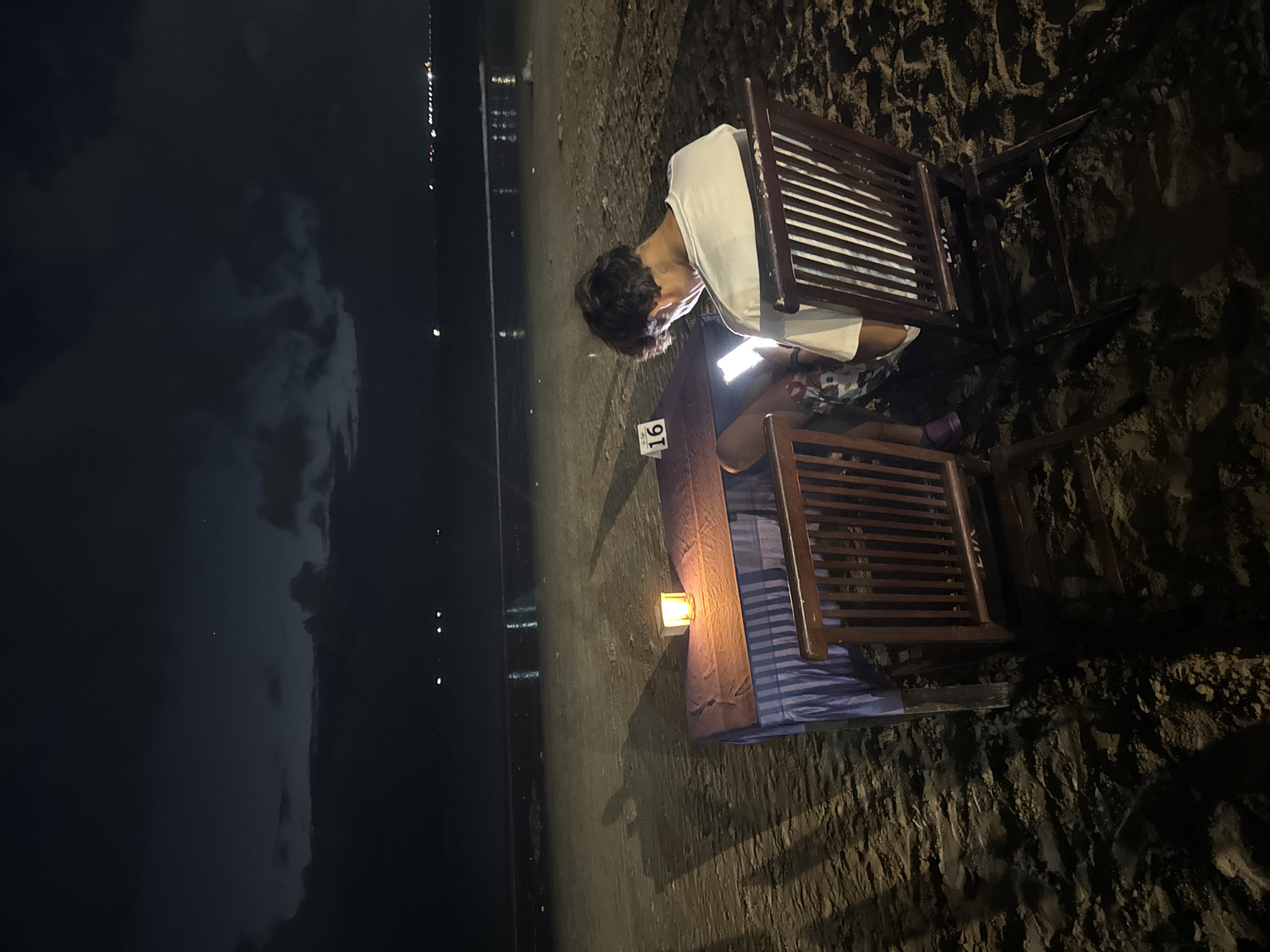 The image depicts a person sitting at a table on a beach at night. The subject is a young person with dark hair, wearing a white t-shirt and patterned shorts. They are hunched over a lit-up mobile phone, engrossed in its screen. The person is seated at a table covered with a blue and white striped cloth, with a small lantern providing a warm glow. Two wooden chairs with slatted backs are visible, one occupied by the person and another next to them. The table has a sign with the number "16" on it.
The setting is a beach, indicated by the sandy ground with visible textures and debris. The background shows a dark expanse, likely the sea, with distant lights suggesting a town or resorts along the coastline. The sky is dark with some clouds, hinting at nighttime. The overall mood is quiet and solitary, with the person focused on their device amidst the natural setting. The presence of the lantern and chairs suggests this might be an outdoor seating area of a restaurant or cafe on the beach, possibly in Kuta Selatan, Indonesia, as suggested by the location context. The time of day is clearly night, and the weather appears calm.