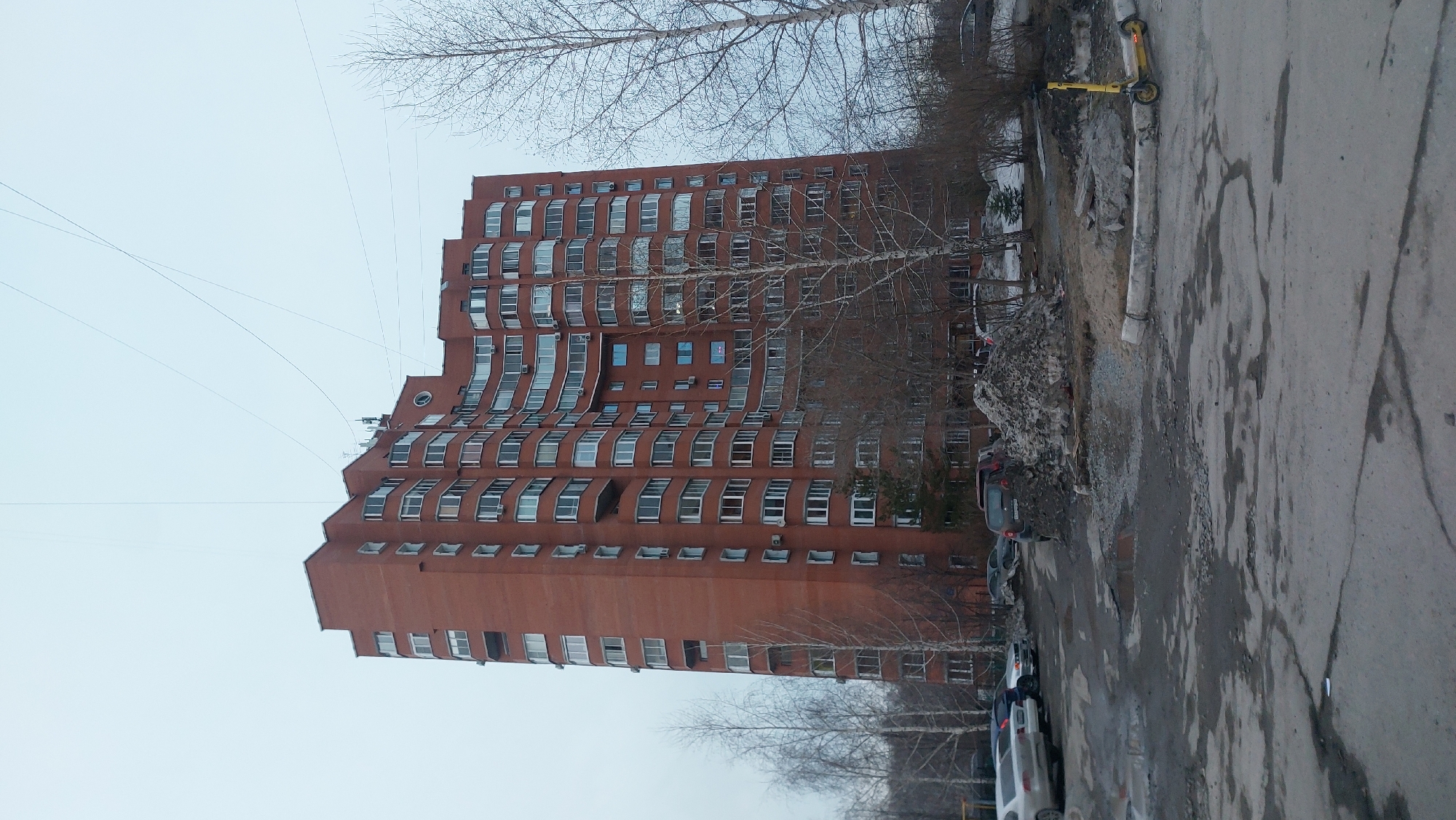 The image depicts a tall, red brick apartment building in Novosibirsk, Russia. The building has a distinctive, curved facade and numerous windows. Bare trees surround the building, indicating it is likely winter or early spring. Several parked cars line the street in front of the building. The pavement appears cracked and in disrepair, with evidence of construction debris. A yellow scooter is standing in the top right of the image. The sky is overcast and gray. Power lines run across the sky near the top of the building.