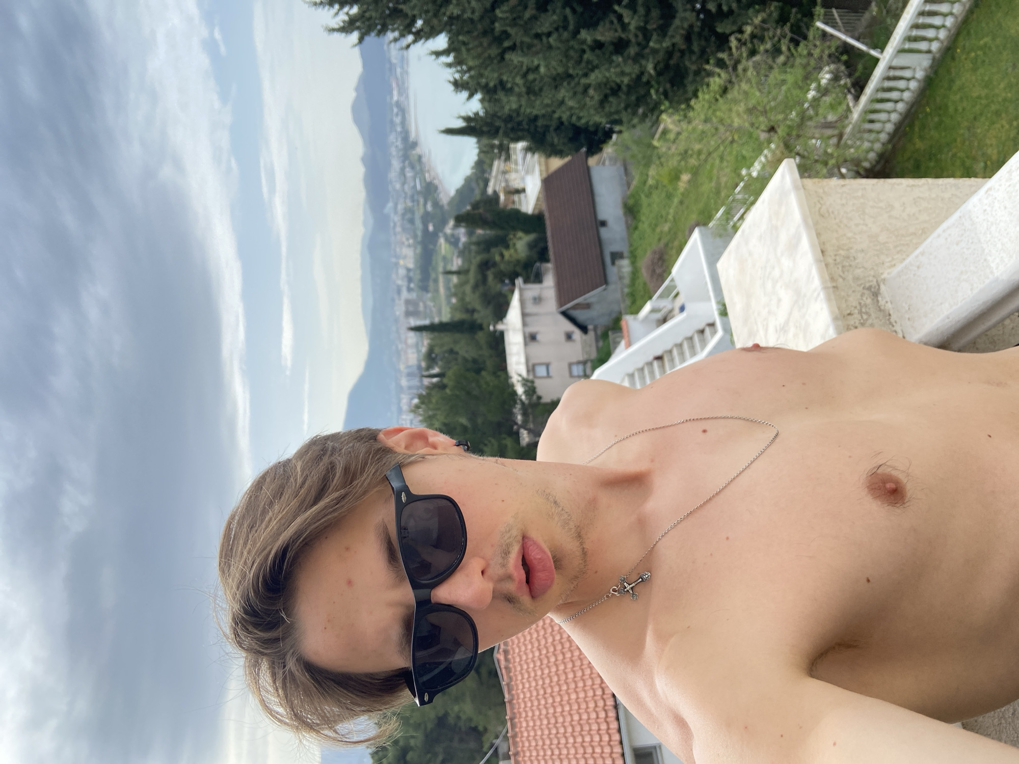 A shirtless young man wearing sunglasses and a silver cross necklace poses for a selfie on a balcony in Šušanj, Montenegro. The backdrop includes buildings, trees, mountains, a coastline, and a partly cloudy sky. The photo appears to be taken on a residential balcony, with railing visible on the right. The city of Šušanj is visible in the background.