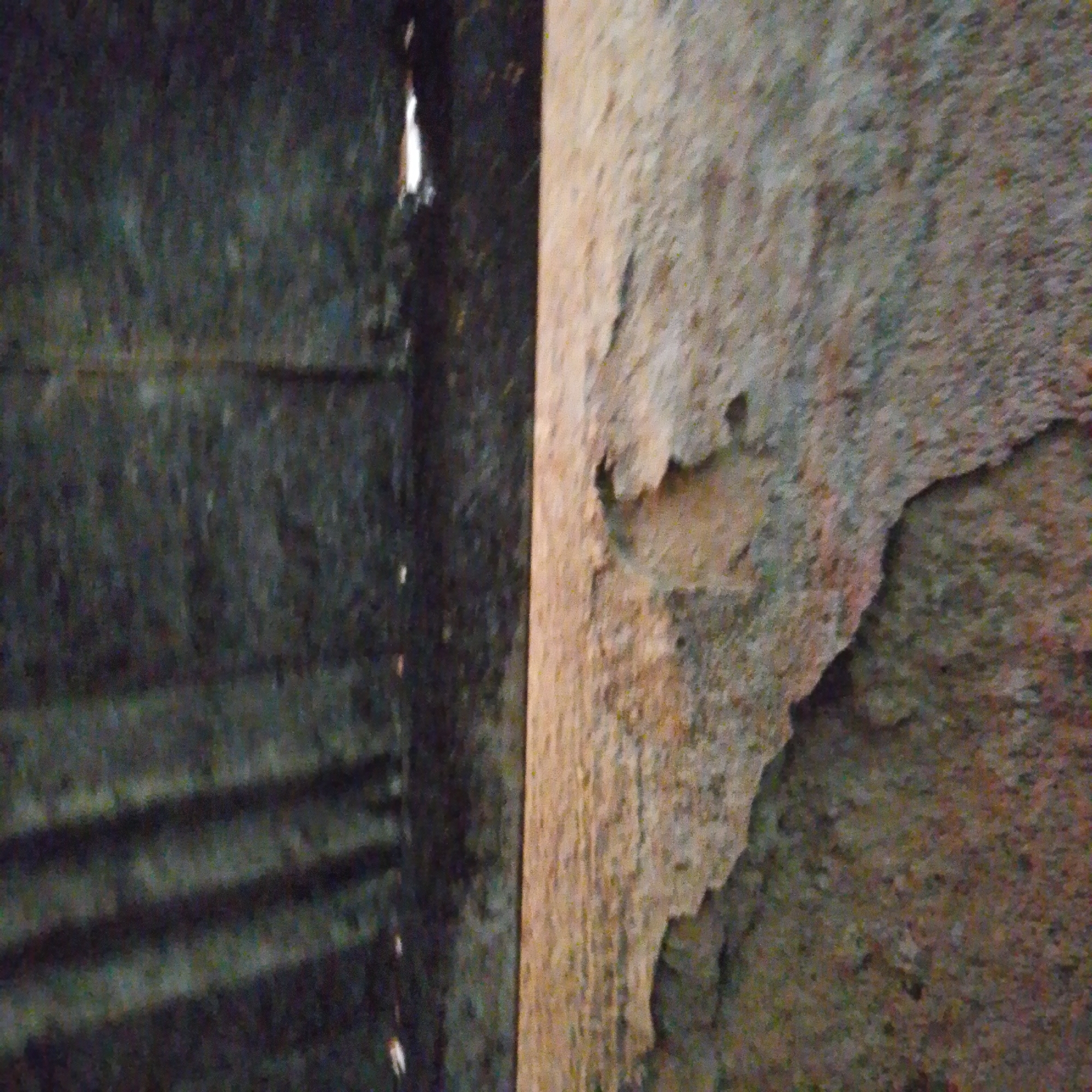 This image captures a close-up view of a textured wall, likely made of concrete or a similar material, with a distinct vertical division. On the left side, the wall appears darker and exhibits horizontal lines, possibly indicating construction seams or markings. There is a narrow vertical gap or opening on the far left, through which a bright white light shines. Steps are faintly visible leading downwards on the left side. The right side of the wall is lighter in color and displays a rough, uneven texture with chipped or peeling sections. The lighting suggests an indoor environment, possibly a dimly lit corridor or the interior of a structure in Jalingo, Nigeria.