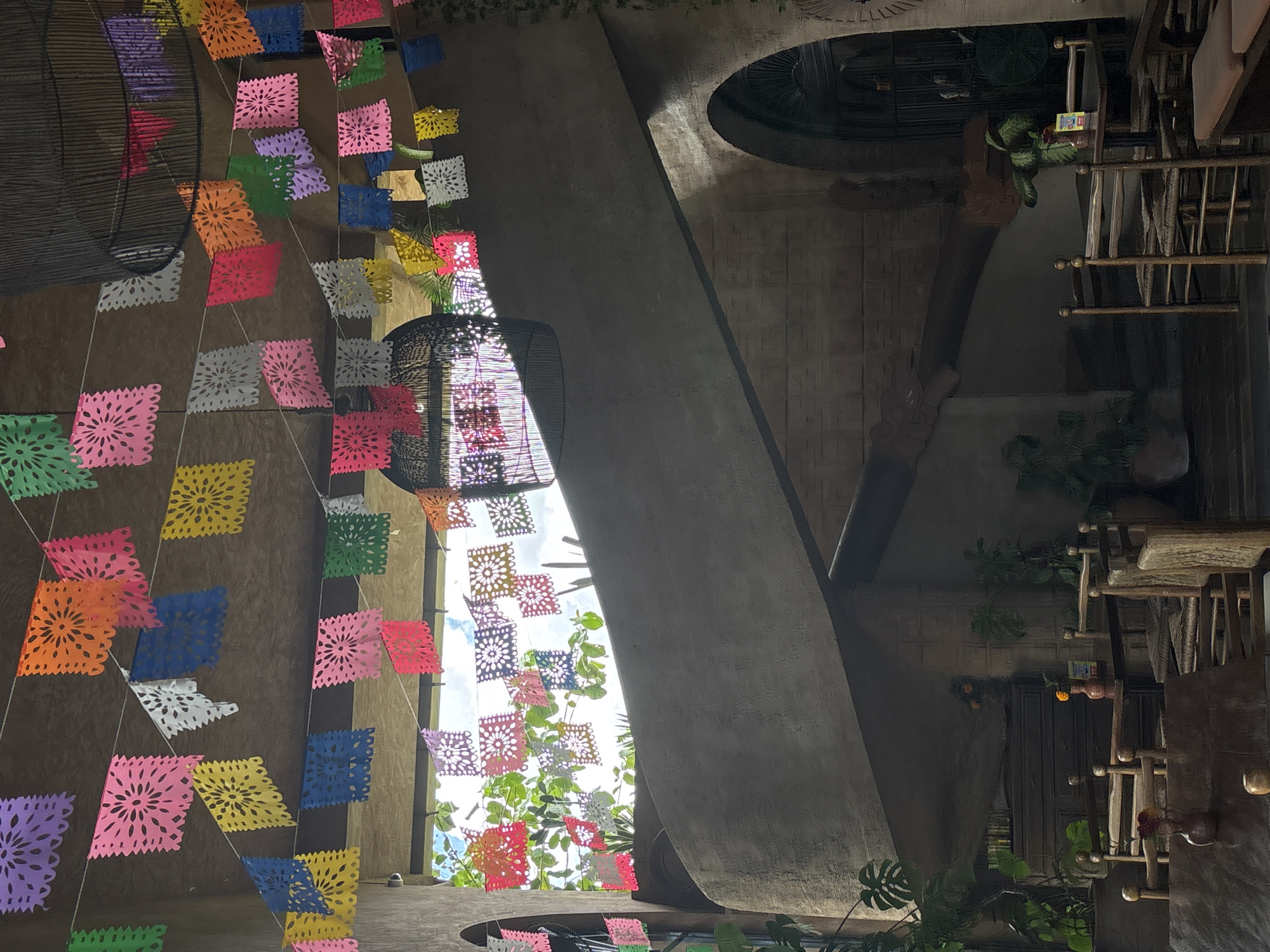 The image depicts an indoor courtyard or atrium in a building, decorated with colorful papel picado banners strung across the space. A large concrete architectural element, possibly a beam or wall section, diagonally bisects the view. Daylight streams through an opening, illuminating foliage and the sky above. An intricate lantern hangs from the ceiling. In the background, elements of a building structure are visible, including what appear to be tables and other furnishings. The scene is reminiscent of a building located in Mengwi, Indonesia.