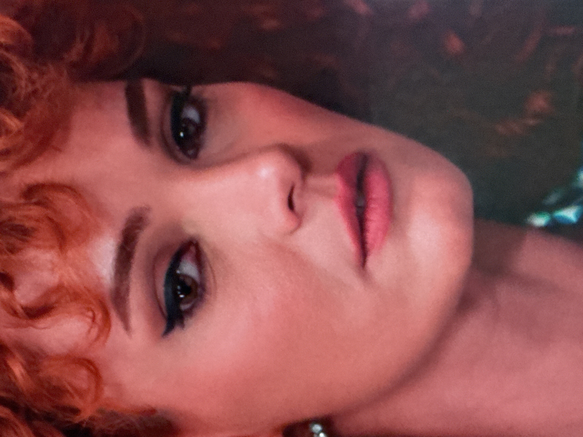 This is a close-up, headshot of a woman with bright red, curly hair. She has fair skin with a pinkish tone, dark eyeliner creating a winged effect, and pink lipstick. A small, shimmering earring is visible on her left ear. Her gaze is directed slightly to the left of the camera. The background is dark and out of focus, with hints of her curly hair extending to the right. The image appears to be a still from a film or a portrait, with a soft, warm lighting that emphasizes the subject's features. The specific location in Uyutnoye, Ukraine cannot be determined from the provided image.