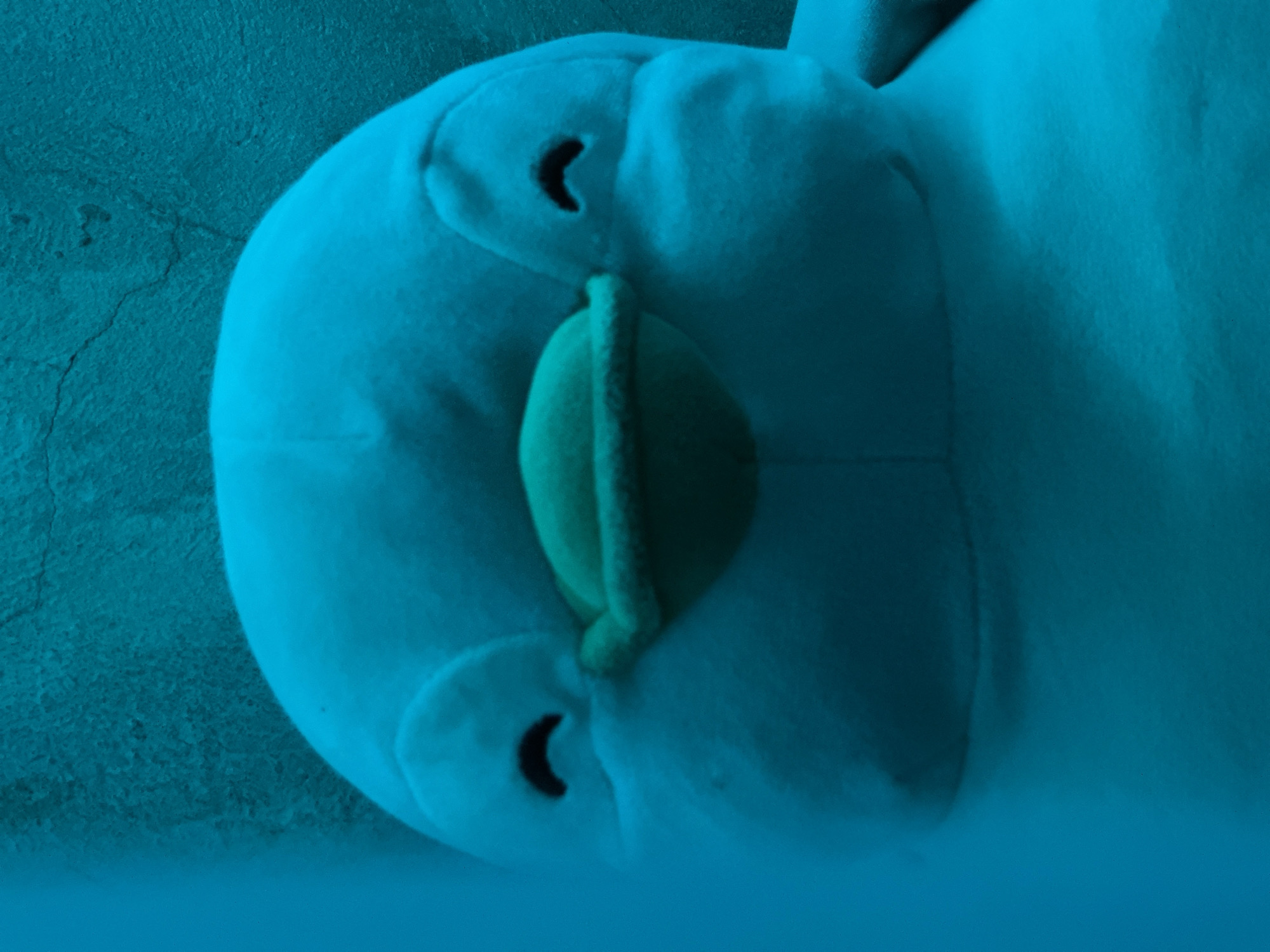 The image is a close-up, head-on shot of a blue plush toy, likely a penguin or a similar cartoon character. The toy has a rounded head, two dark, crescent-shaped eyes, and a yellowish-green beak or mouth area. The background appears to be a textured, light-colored wall, possibly concrete, with some visible cracks. The lighting is dim, casting a predominantly blue hue over the entire scene, with some areas of shadow and lighter blue. No people or specific Davao City landmarks are visible. The setting appears to be indoors.