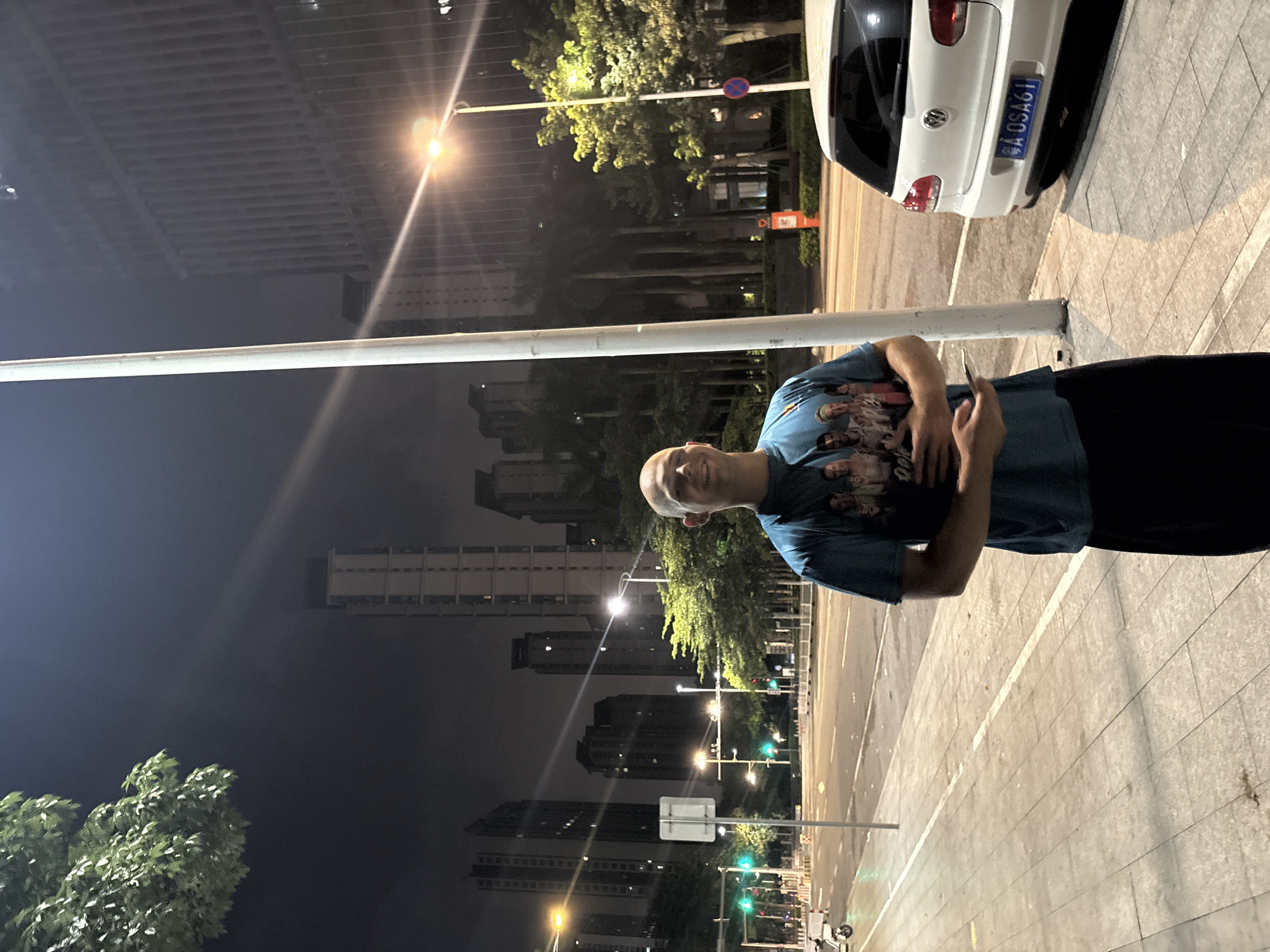 A man stands on a sidewalk at night in Haizhu District, China, holding a smartphone. He is wearing a teal t-shirt with a graphic print on the front and black pants. The man has a shaved head and is looking towards the camera with a slight smile. In the background, a white Volkswagen car is parked on the street, which is illuminated by streetlights and the lights from surrounding buildings. The scene includes a traffic light, trees, and tall buildings, indicating an urban environment.