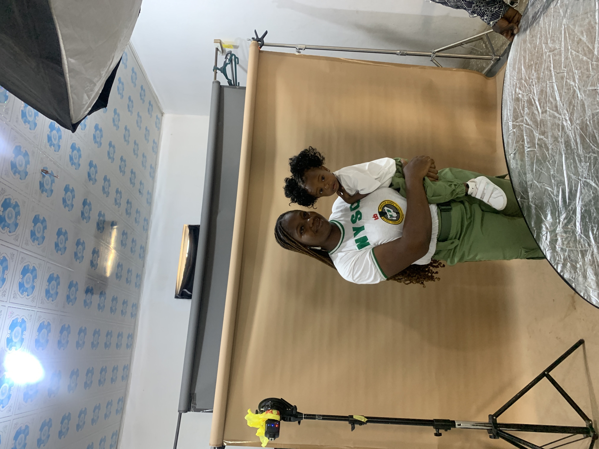 A woman and a child are in a photography studio setting. The woman is wearing a white t-shirt with green trim and an NYSC (National Youth Service Corps) logo, along with green pants. She's holding the child, who is wearing a white shirt and green pants. They are standing in front of a solid brown backdrop. Photography equipment is visible, including softboxes and a reflector. The location appears to be indoors, as suggested by the tiled wall. The tiled walls are blue and white. Gura Top, Nigeria cannot be verified as the specific location for this photograph.