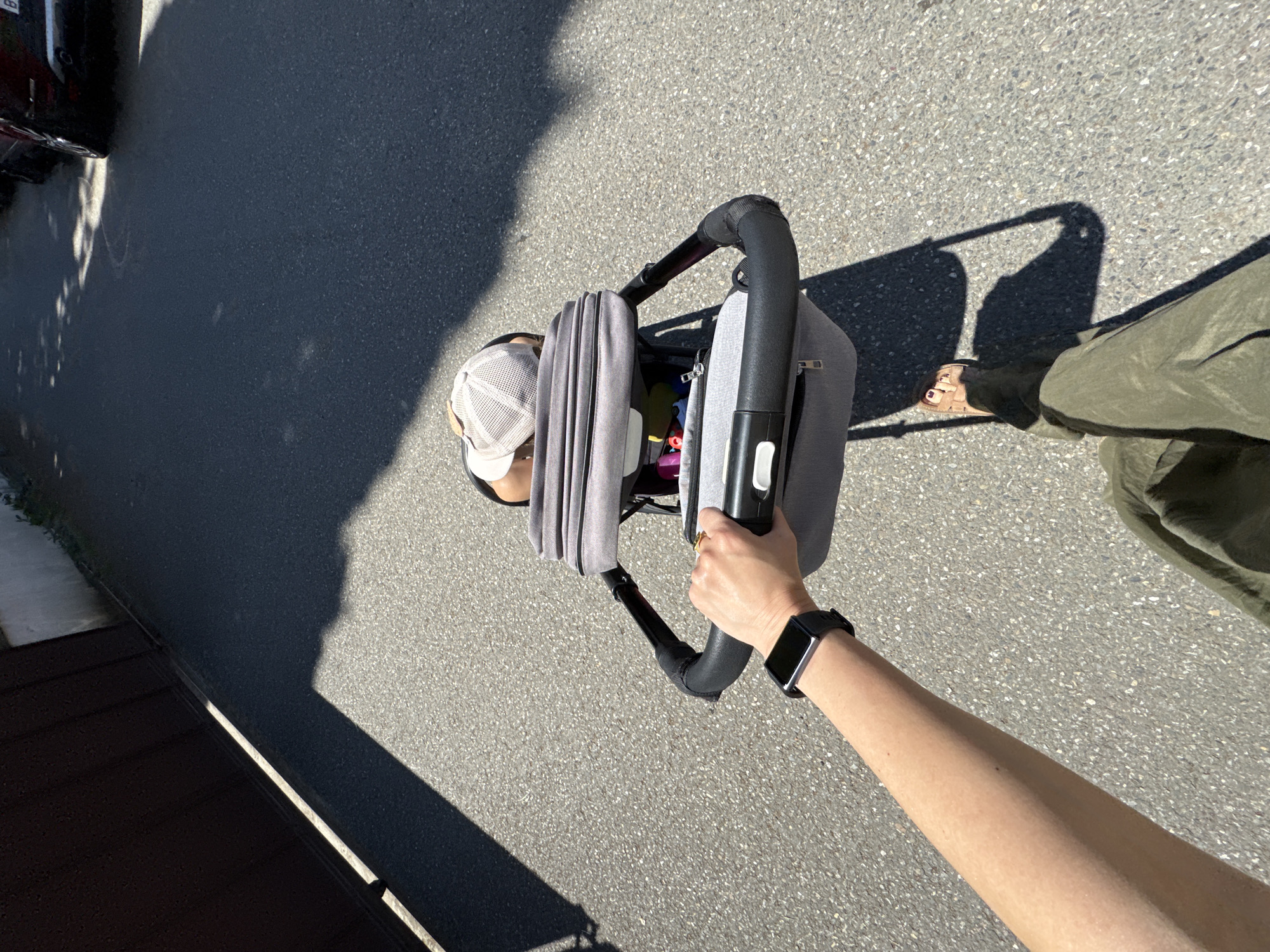An overhead, high-angle shot captures a person pushing a stroller on an asphalt path under bright sunlight in Sochi, Russia. The perspective is from directly above the person's right arm, which extends into the frame from the lower right, holding the black handle of a grey stroller. A black smartwatch is visible on the person's wrist, and a gold ring is on their finger.
A small child or baby, wearing a light-colored mesh cap, is partially visible inside the grey stroller. On the ground, the person's right foot, adorned with a light-colored sandal and red painted toenails, is visible, along with part of an olive green pant leg.
The asphalt ground is mottled with small stones. Strong, dark shadows of trees or foliage, as well as the person and the stroller, are cast across the path, indicating clear, sunny daytime weather, likely late morning or early afternoon. In the upper left corner, the dark chassis of a parked car is partially visible, with a faint white letter 'B' discernable on its surface. A low wall or curb runs along the bottom left edge of the frame.
