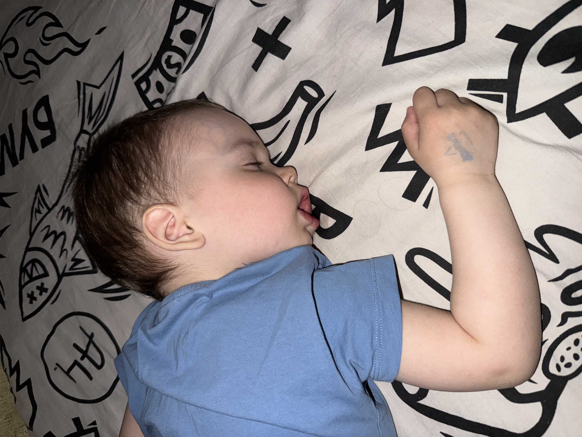 A young child, possibly a toddler, is lying on their side on a bed. They are wearing a blue t-shirt. The child's mouth is slightly open, and their tongue is visible. The child's right arm is raised with the hand near their head, and there appears to be a faint blue mark on the back of the hand. The bed is covered in a white sheet with black graphic designs. The location is unknown and there is no indication that this image was taken in Rusanovo, Russia.
