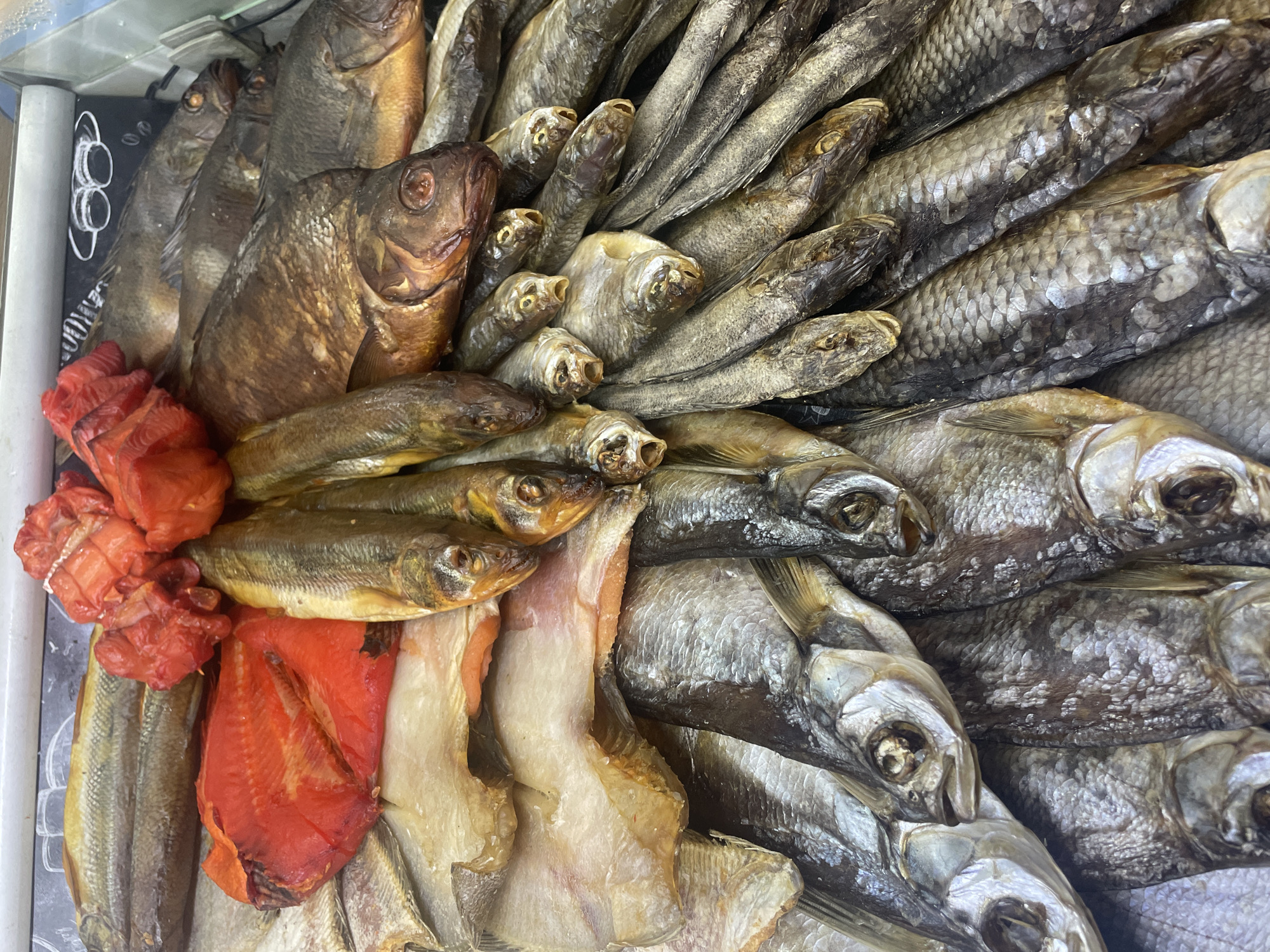 The image depicts a close-up, overhead view of a display of dried and smoked fish. The fish vary in size and color, with some being a light silver or tan, while others are a darker brown or golden hue. Some pieces appear to be whole, while others are filleted or cut. In the top center of the image, there are several bright red pieces of what appears to be smoked salmon. The fish are arranged in a dense pile, filling the frame. The background is out of focus, but appears to be a white or light-colored surface, possibly a counter or display case. There are no people visible in the image, and no immediate indication of the specific location within the city of Zhukovo, Russia. The scene is a still life focusing on the presentation of food products.