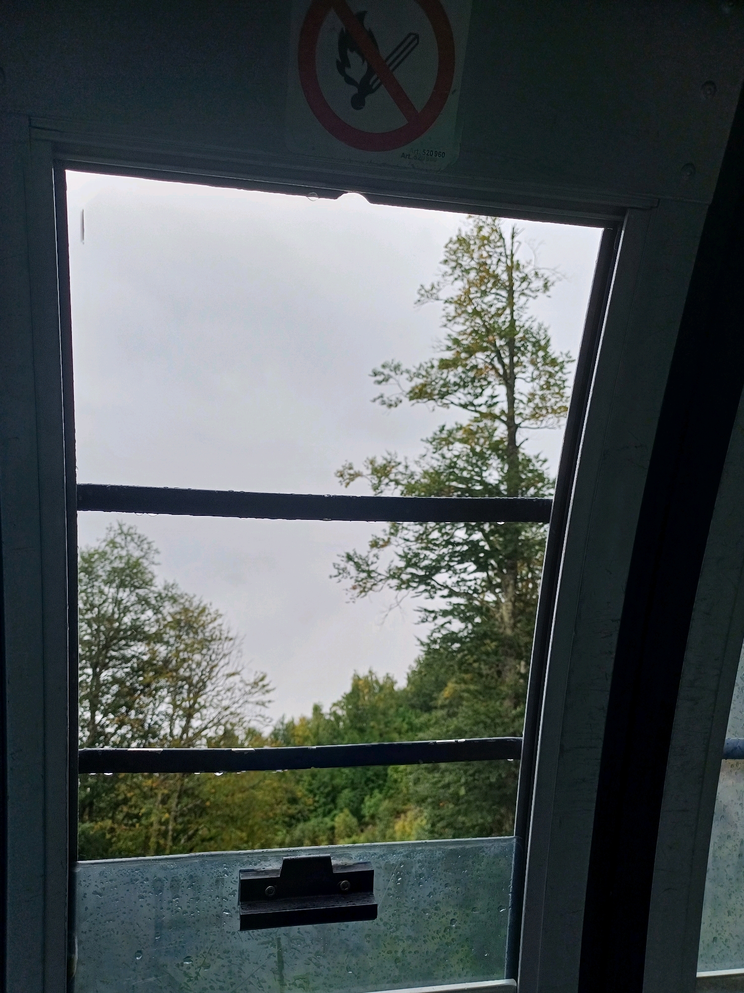 This image shows a view through a window from inside what appears to be a cabin, possibly a cable car, looking out at a forest on a cloudy day. The window is slightly wet with raindrops visible on the glass and along the horizontal bar that crosses the view. A circular warning sign with a red border and a diagonal line through a symbol of a person with a cigarette is visible at the top of the frame. The trees outside show autumn colors, with some green and yellow foliage. There is a dark, curved structure on the right side of the frame, which is part of the cabin's interior. A latch mechanism is visible on the lower part of the window. The scene is likely set in Estosadok, Russia, a location known for its mountainous terrain and ski resorts, which often utilize cable cars.