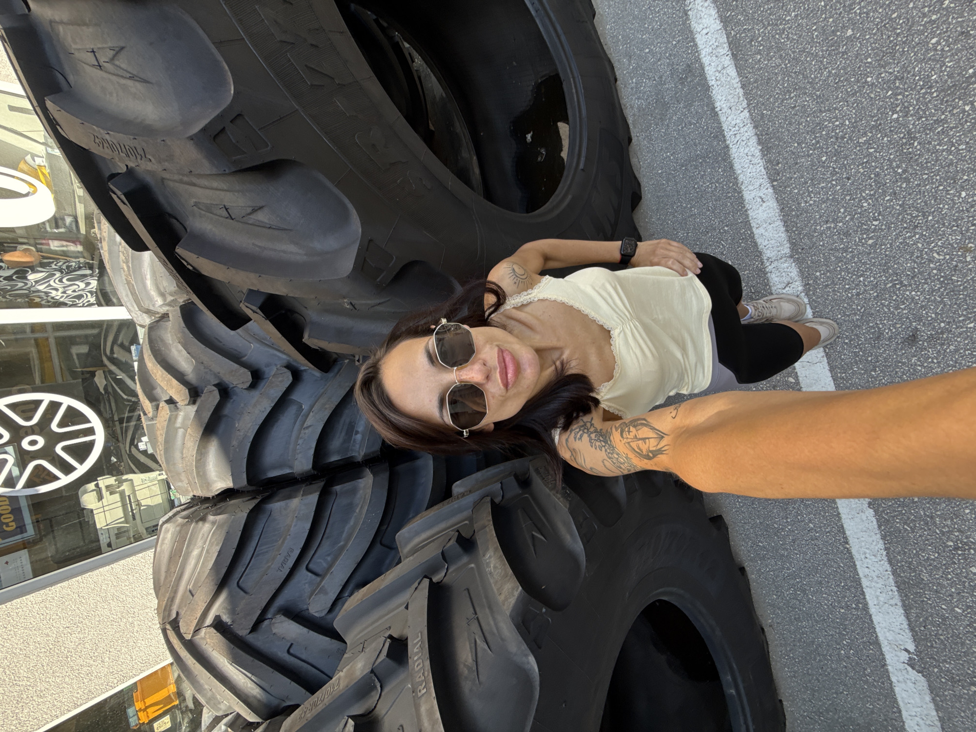 A woman with sunglasses poses next to a stack of large tires outside of a building, possibly a tire shop. The tires are black and appear to be for agricultural or industrial vehicles. The location is Pleven, Bulgaria, based on the storefront and general surroundings. The woman is fair-skinned and wearing a light-colored top and dark leggings. She has tattoos on her arms. The tires are stacked next to a parking space. The area is paved.