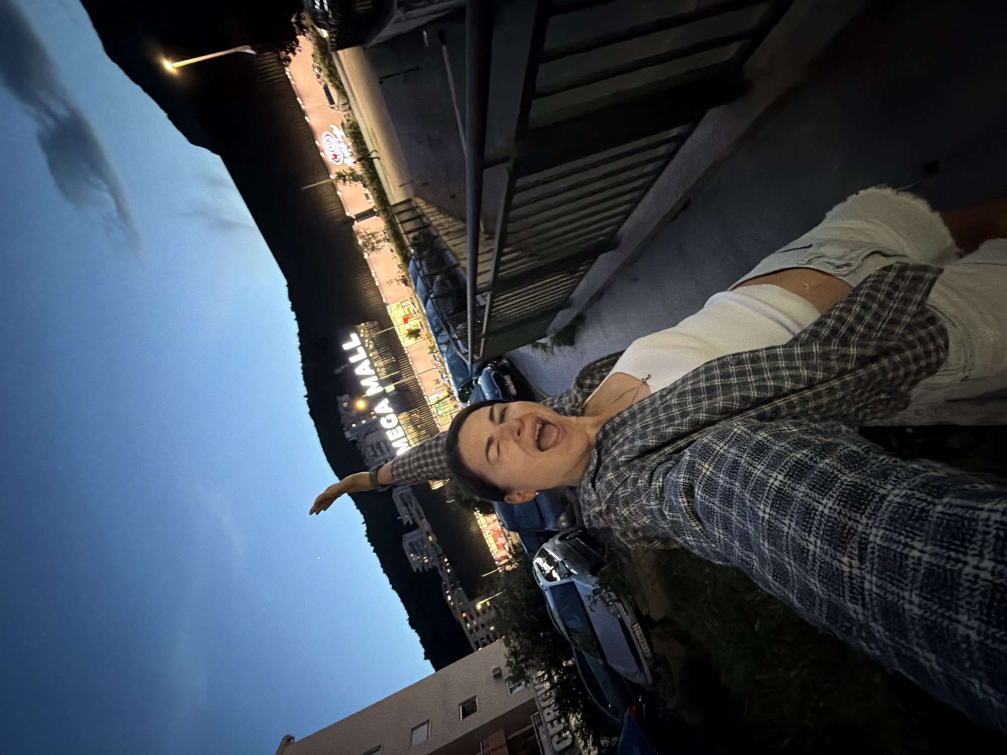 A young woman is captured in an enthusiastic pose against an evening or night backdrop. The image is taken at an unusual angle, making her appear to be lying down or leaning dramatically. She is smiling widely with her mouth open as if laughing or shouting, eyes partially closed, conveying a sense of joy and exuberance. She wears a white top underneath a plaid shirt with blue/grey and white checks, and light-colored shorts or pants. Her left arm is extended outwards, adding to the dynamic nature of her pose.
In the background, a large, illuminated sign brightly displays "MEGA MALL" across a distant hillside or building. Numerous other buildings with illuminated windows and various smaller lights are visible, indicating an active urban or commercial environment. Several cars are parked below, suggesting a street or parking lot. The sky overhead is a deep twilight blue with faint clouds, marking the time as dusk or night. The scene captures a moment of playful energy in Donji Pobori, Montenegro.